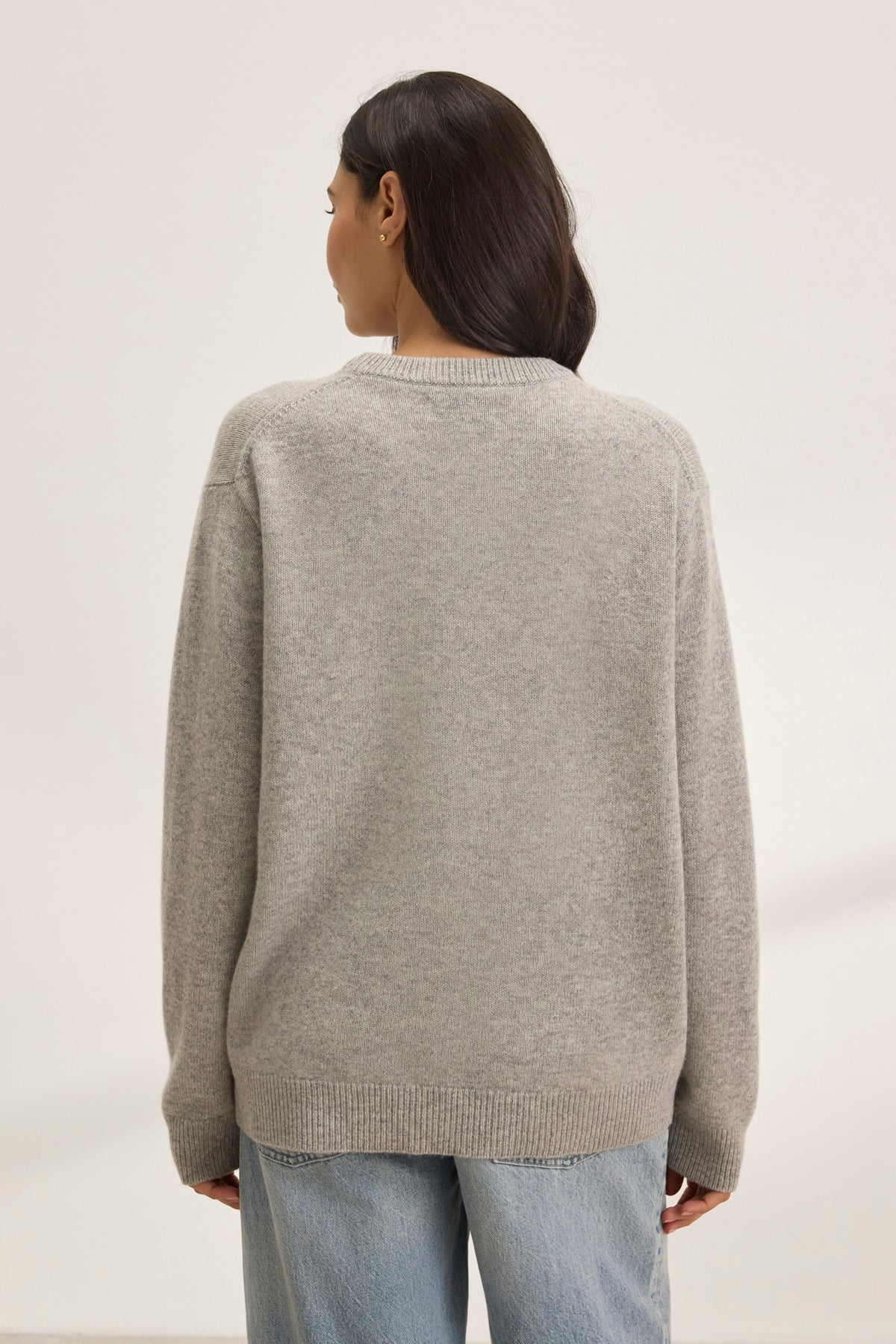   Someone is clad in a cozy, oversized gray STEVIE WOOL CASHMERE SWEATER by Velvet by Graham & Spencer. They pair it with blue jeans and stand facing away against a plain background. 