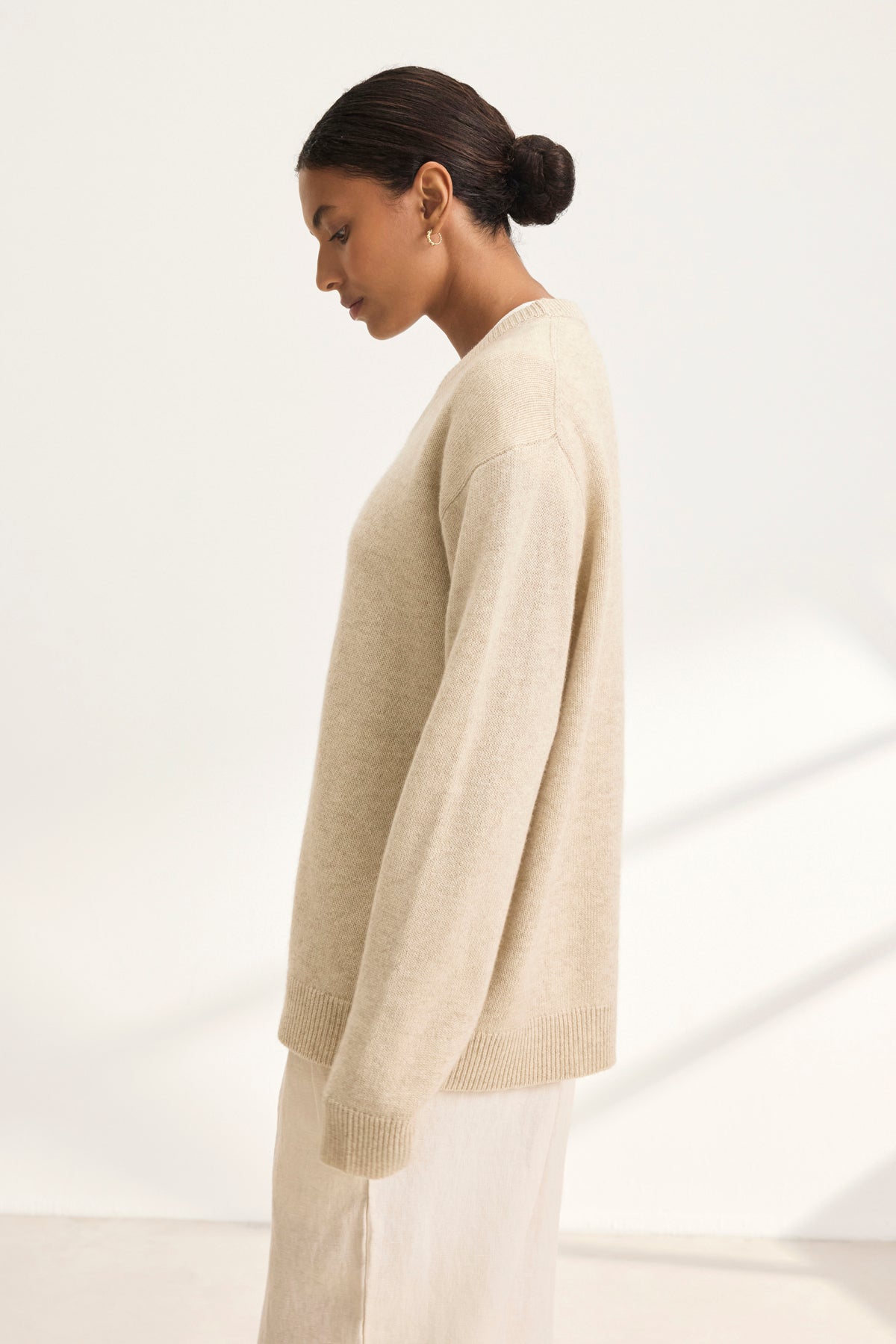   A person with dark hair in a bun is wearing the oversized STEVIE WOOL CASHMERE SWEATER by Velvet by Graham & Spencer and a light skirt, standing sideways against a light background. 