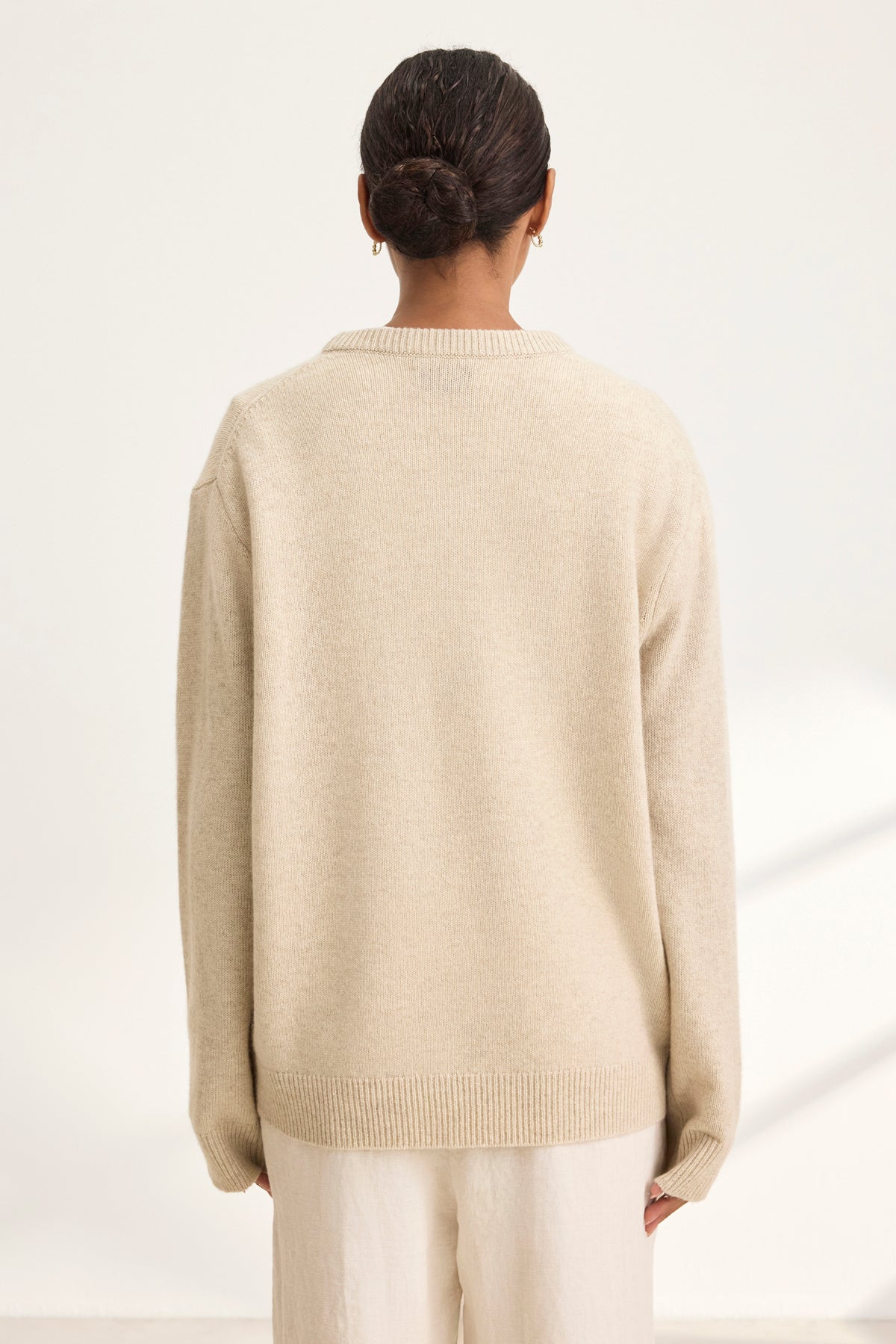   Wearing a Velvet by Graham & Spencer STEVIE WOOL CASHMERE SWEATER with cream pants, a person stands against a light background. Their hair is tied in a bun, enhancing the cozy and relaxed vibe. 