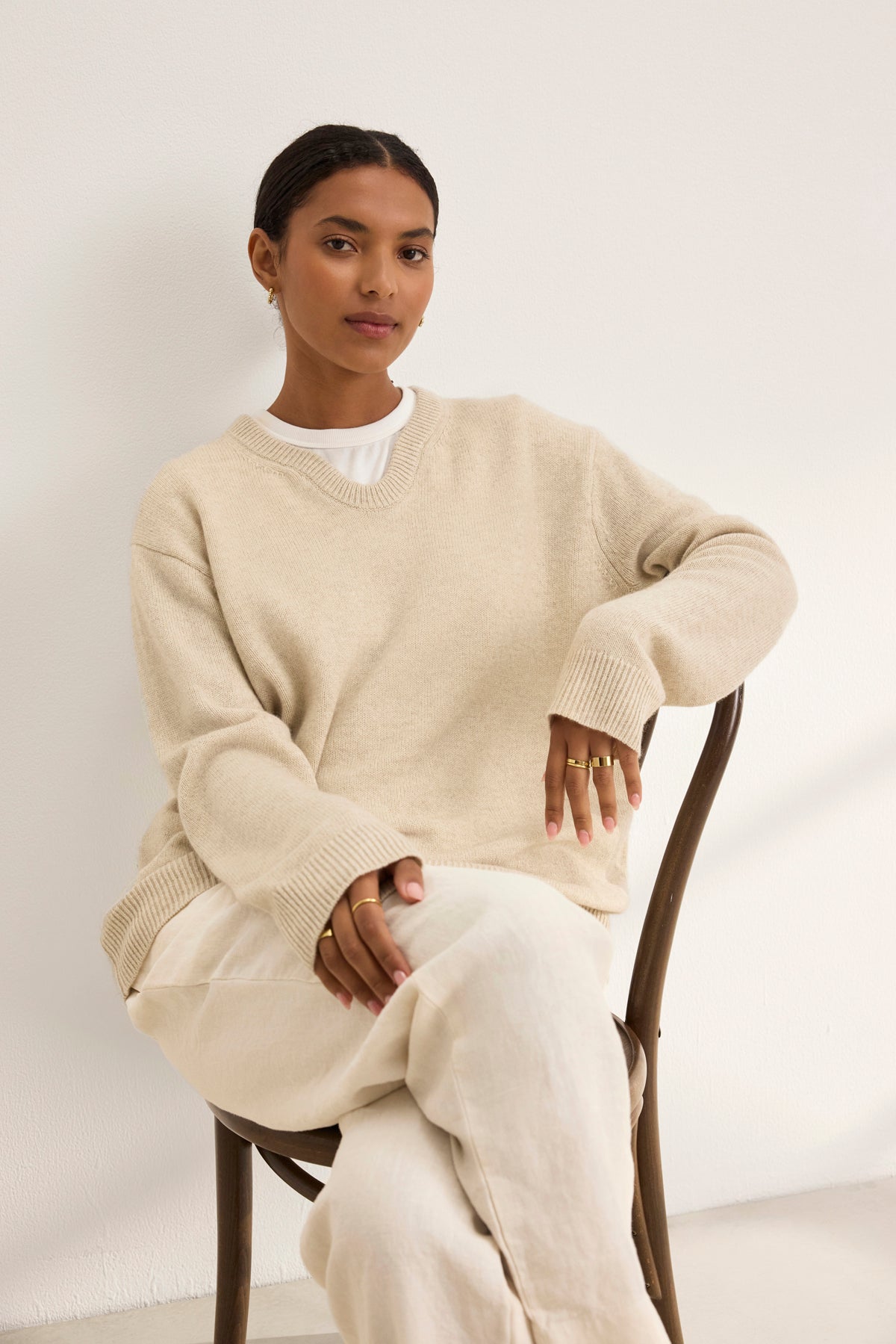   A person sits on a chair with hands on their lap, wearing the STEVIE WOOL CASHMERE SWEATER by Velvet by Graham & Spencer and light pants, against a plain white background. 