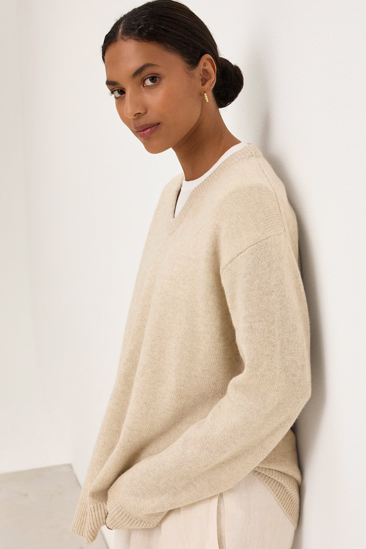   A person wearing the STEVIE WOOL CASHMERE SWEATER by Velvet by Graham & Spencer and white pants stands against a white wall, looking at the camera. 