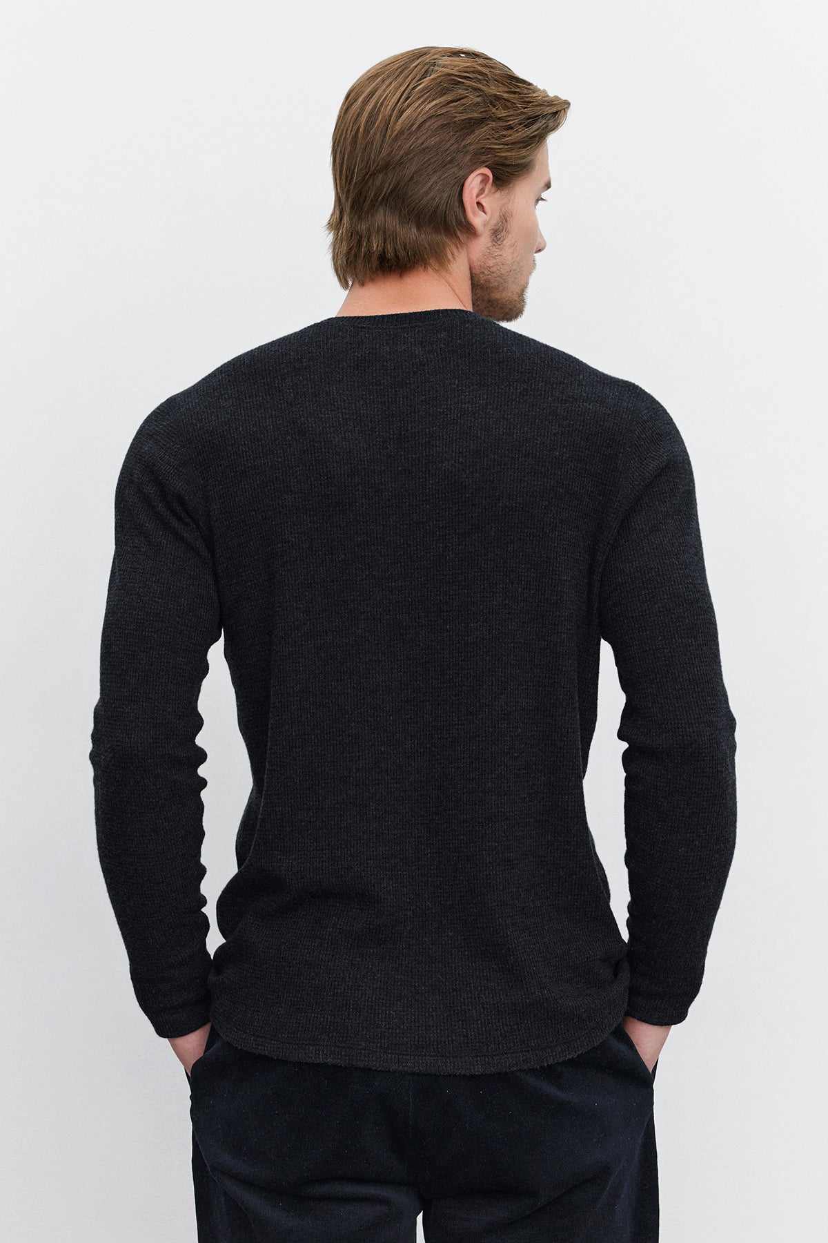   A person with light brown hair stands facing away, wearing a Velvet by Graham & Spencer AMBROSE HENLEY shirt in dark gray with a three-button placket and black pants, against a plain white background. 