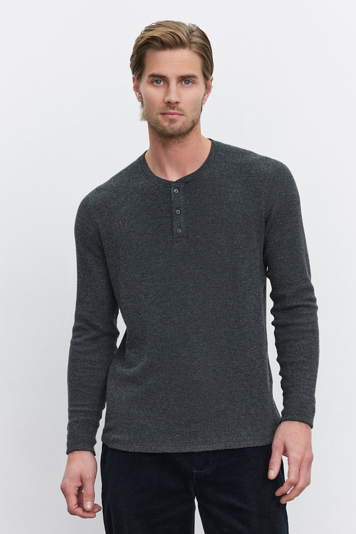   A man with light brown hair and beard is wearing the AMBROSE HENLEY, a dark gray henley with a three-button placket from Velvet by Graham & Spencer, as he stands in front of a plain white background. 