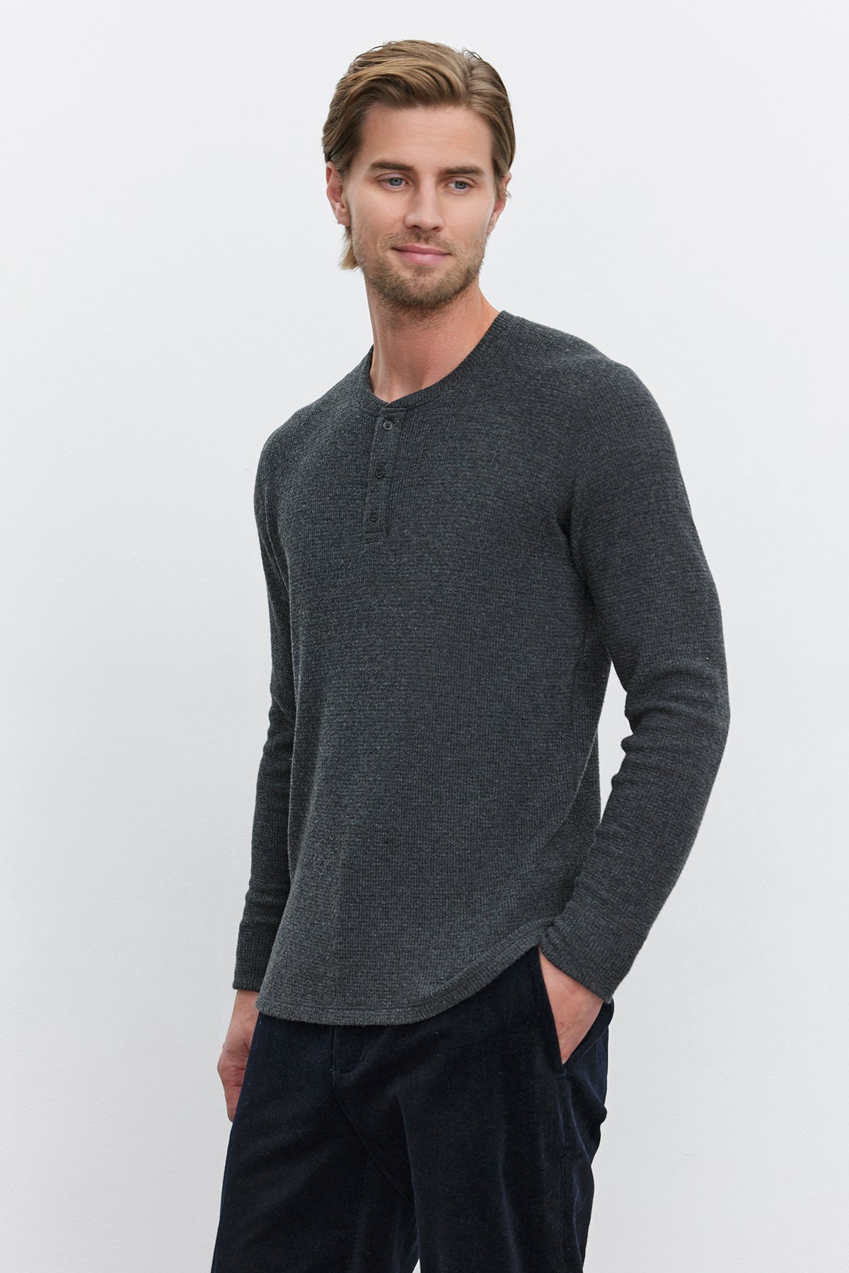   A man with light brown hair stands against a plain white background, wearing the AMBROSE HENLEY by Velvet by Graham & Spencer. The long-sleeve henley shirt in dark gray is made from cozy thermal fabric and paired with dark pants. The three-button placket adds a touch of style as he has his hands in his pockets. 