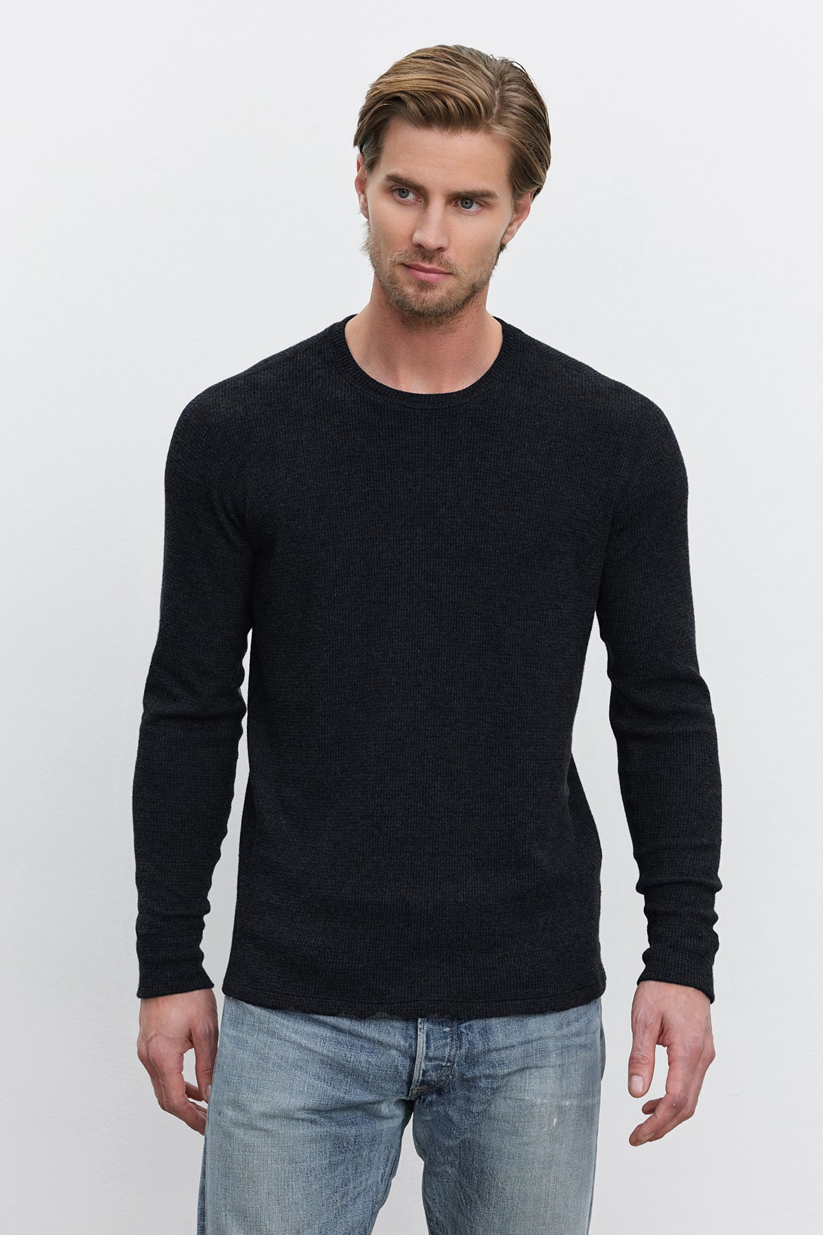   A man with short, light brown hair and a beard is wearing the TOBIAS CREW black long-sleeve sweater by Velvet by Graham & Spencer, made from cozy thermal fabric, along with light blue jeans. He is standing against a plain white background, looking slightly to his right. 