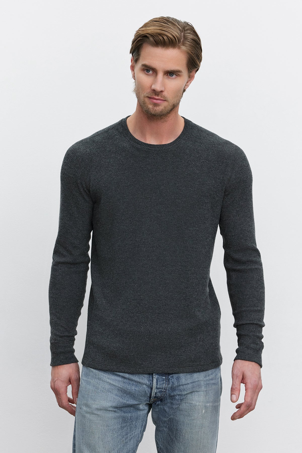   A man with short hair is wearing a TOBIAS CREW from Velvet by Graham & Spencer, a fitted, long-sleeve gray sweater made from triblend cozy thermal fabric, paired with light blue jeans. He is standing against a plain white background. 