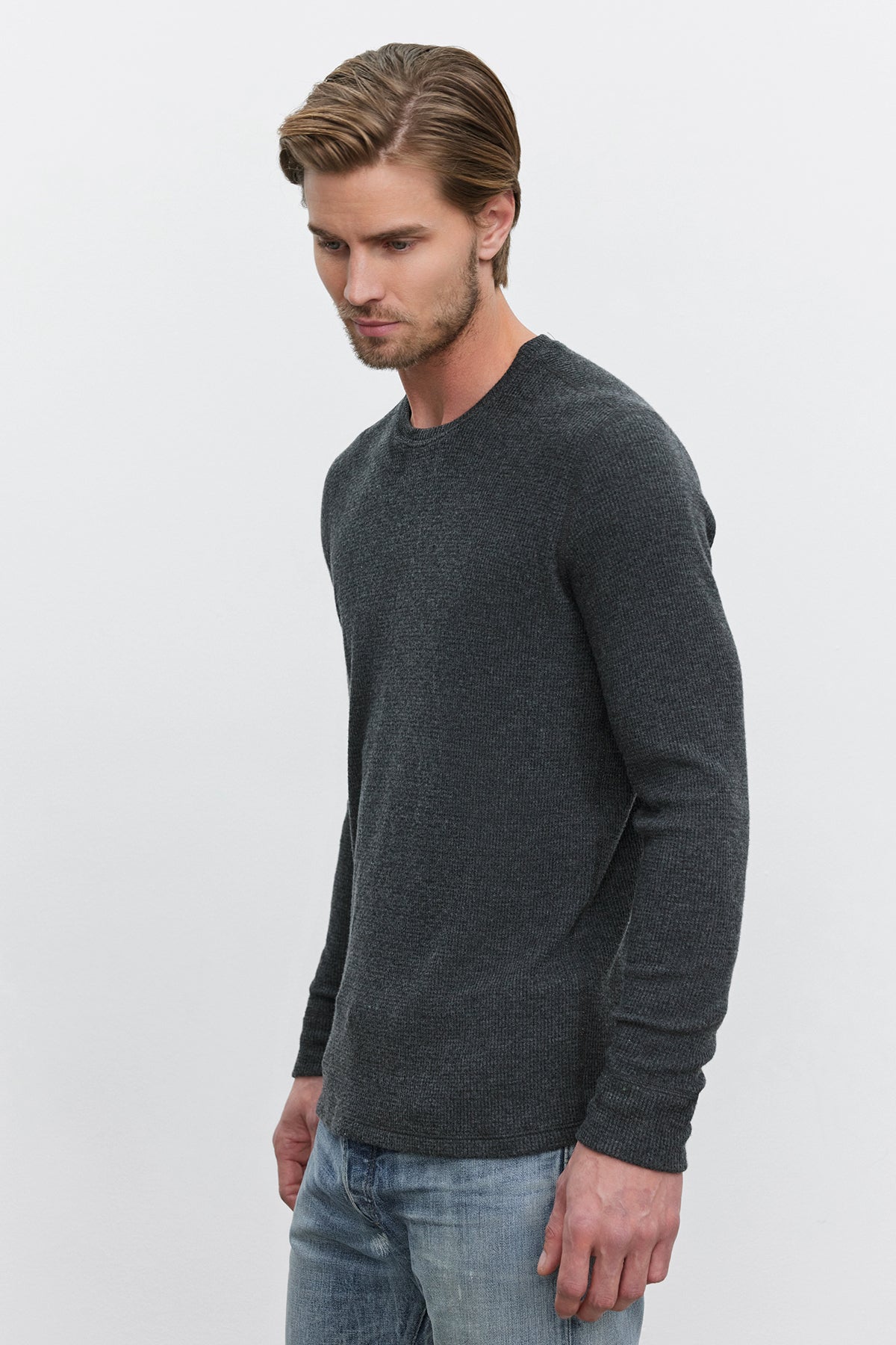   A man with light brown hair, wearing a dark gray TOBIAS CREW long-sleeve shirt made from triblend cozy thermal fabric by Velvet by Graham & Spencer and blue jeans, stands against a plain white background. 