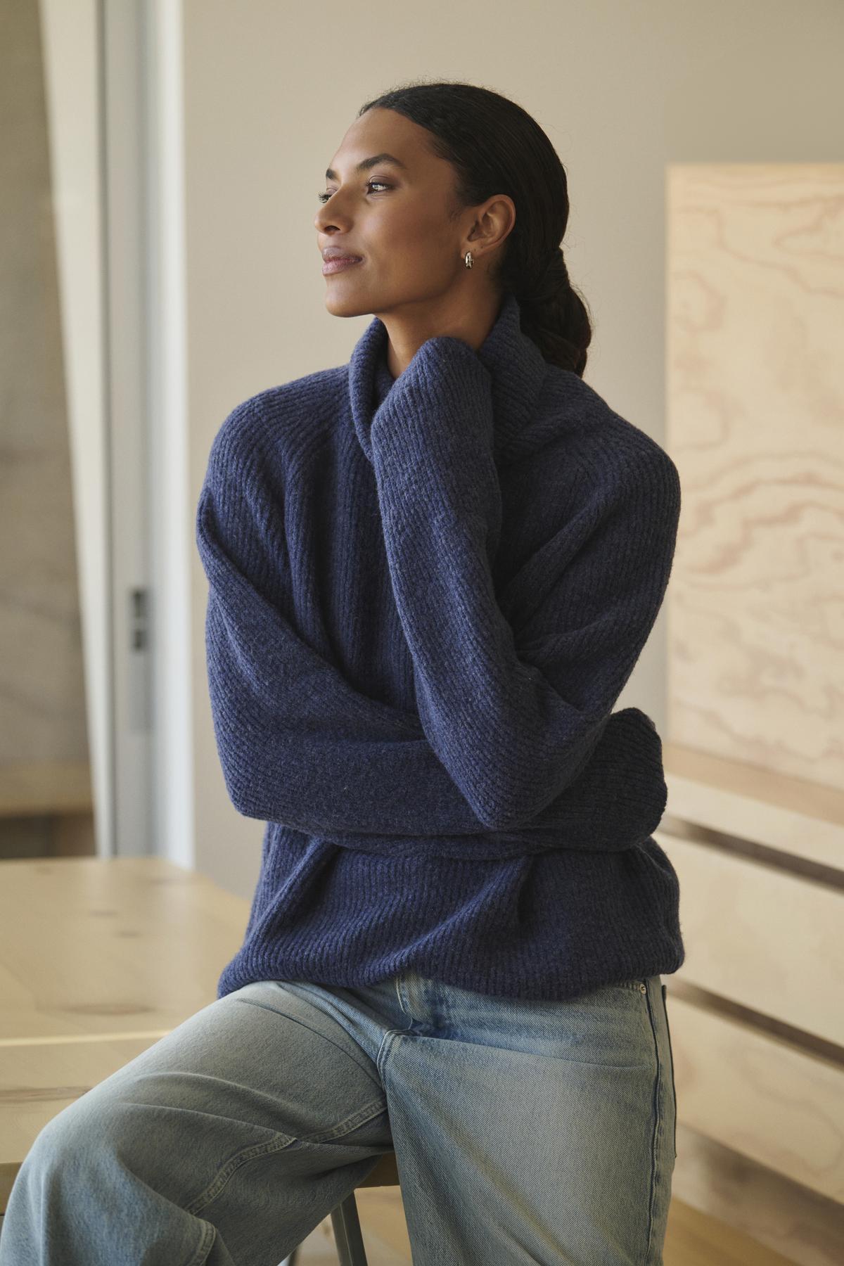 Wearing a cozy JUDITH TURTLENECK SWEATER by Velvet by Graham & Spencer along with jeans, a person sits on a stool, glancing to the side with a neutral expression.-38301587669185