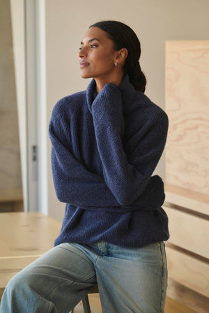 Wearing a cozy JUDITH TURTLENECK SWEATER by Velvet by Graham & Spencer along with jeans, a person sits on a stool, glancing to the side with a neutral expression.