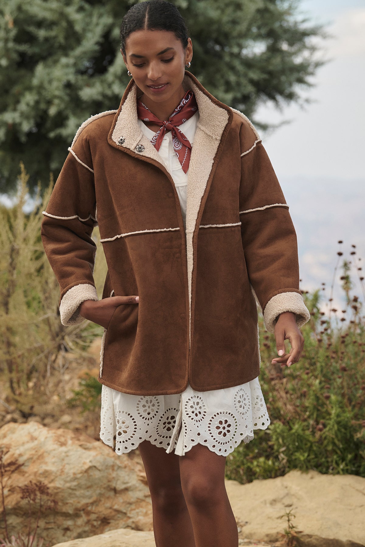 A person wearing the ALBANY LUXE SHERPA REVERSIBLE JACKET by Velvet by Graham & Spencer over a white dress and adorned with a red bandana stands outdoors amid shrubs and rocks, highlighting the eco-friendly faux suede lining that perfectly complements the natural surroundings.-38218871701697