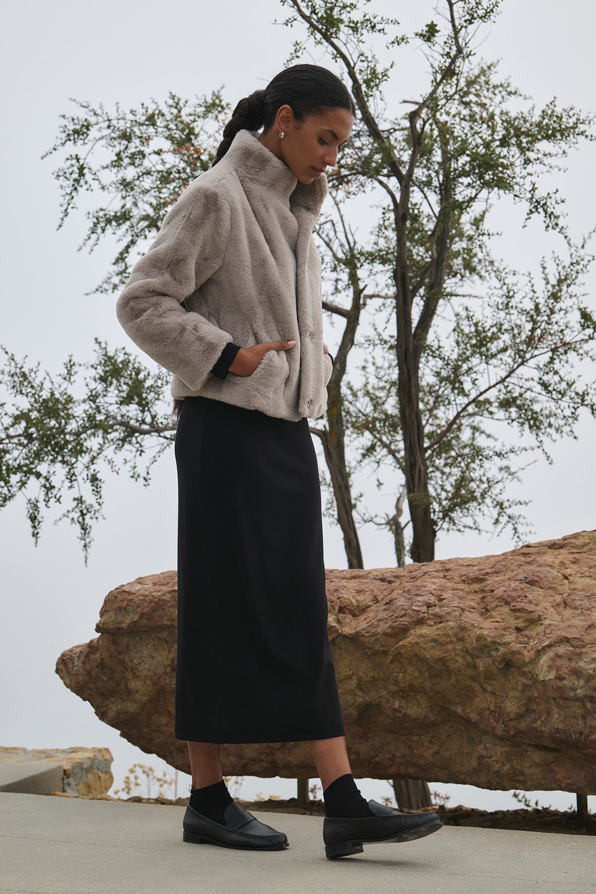   An individual stands outdoors wearing a CORONADO DRESS by Velvet by Jenny Graham, featuring a fitted silhouette and paired with a long black skirt, hands tucked in pockets. A rocky landscape and tree are visible in the background. 