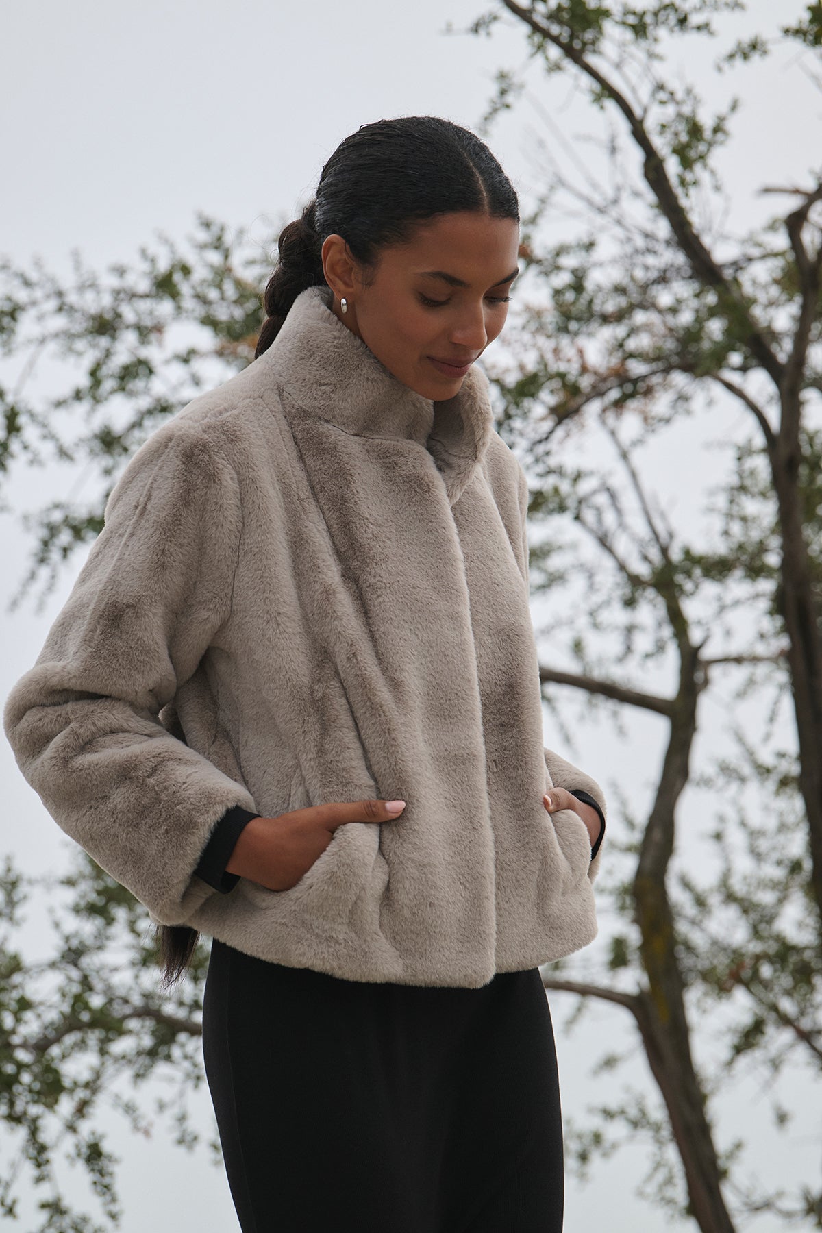   A person stands outdoors with trees in the background, wearing the SHEENA LUXE FAUX FUR JACKET by Velvet by Graham & Spencer, featuring jumbo snap buttons and a light color. 