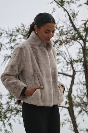 A person stands outdoors with trees in the background, wearing the SHEENA LUXE FAUX FUR JACKET by Velvet by Graham & Spencer, featuring jumbo snap buttons and a light color.