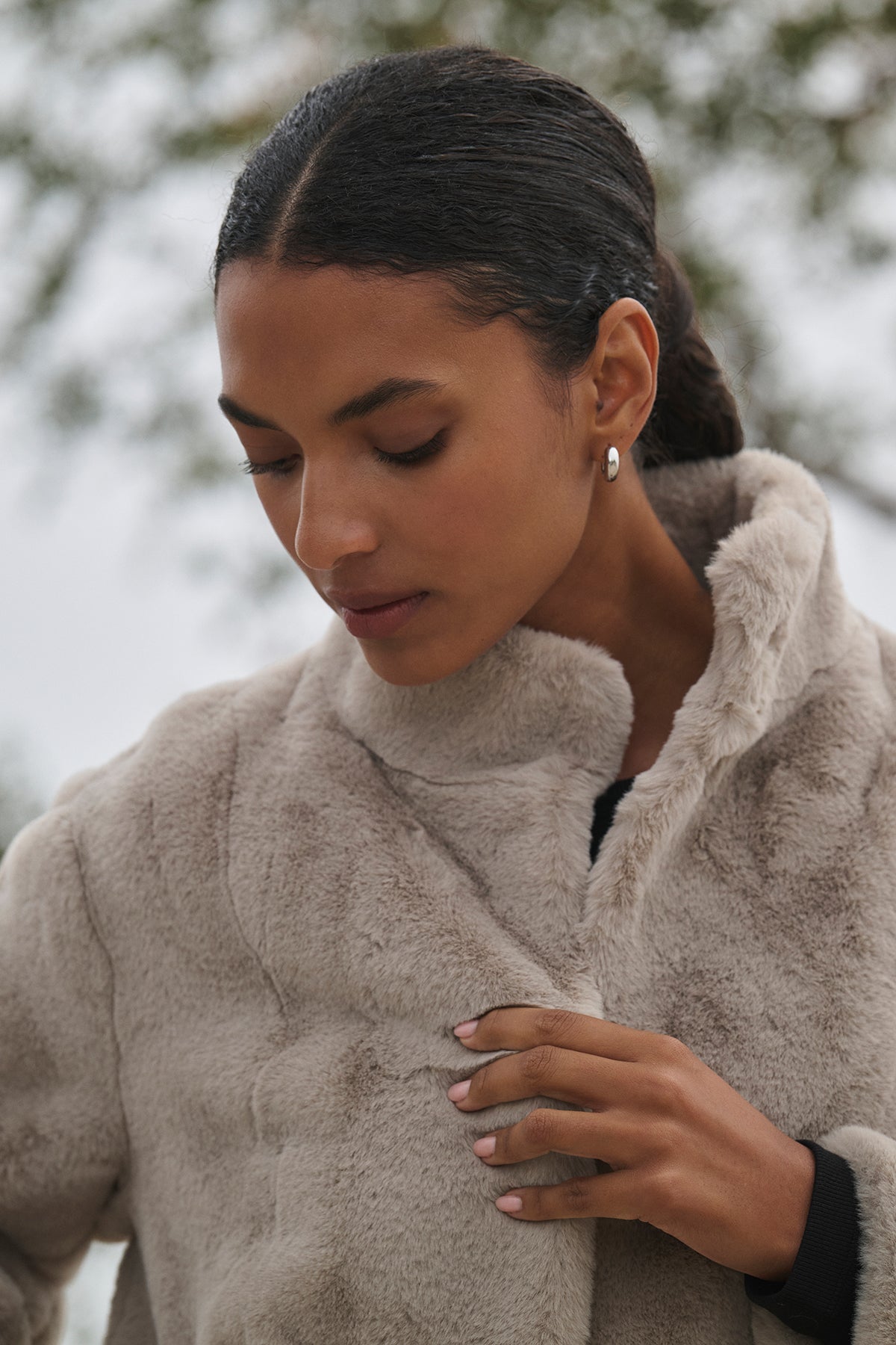 Against a blurred outdoor background, a person with dark hair holds the mock neck collar of a beige SHEENA LUXE FAUX FUR JACKET by Velvet by Graham & Spencer while looking down.-38218839851201