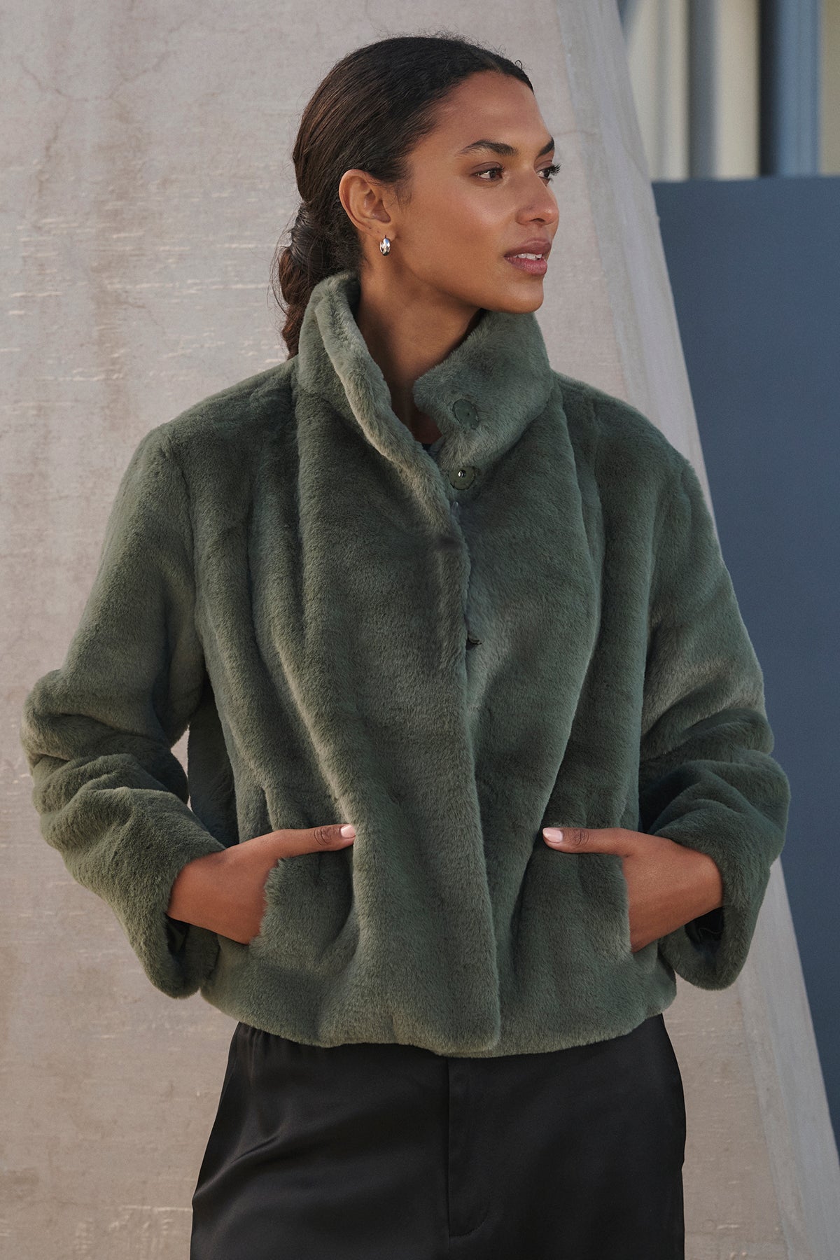   A person wearing the SHEENA LUXE FAUX FUR JACKET from Velvet by Graham & Spencer, featuring a mock neck collar, pairs it stylishly with black pants while standing against a textured wall. 