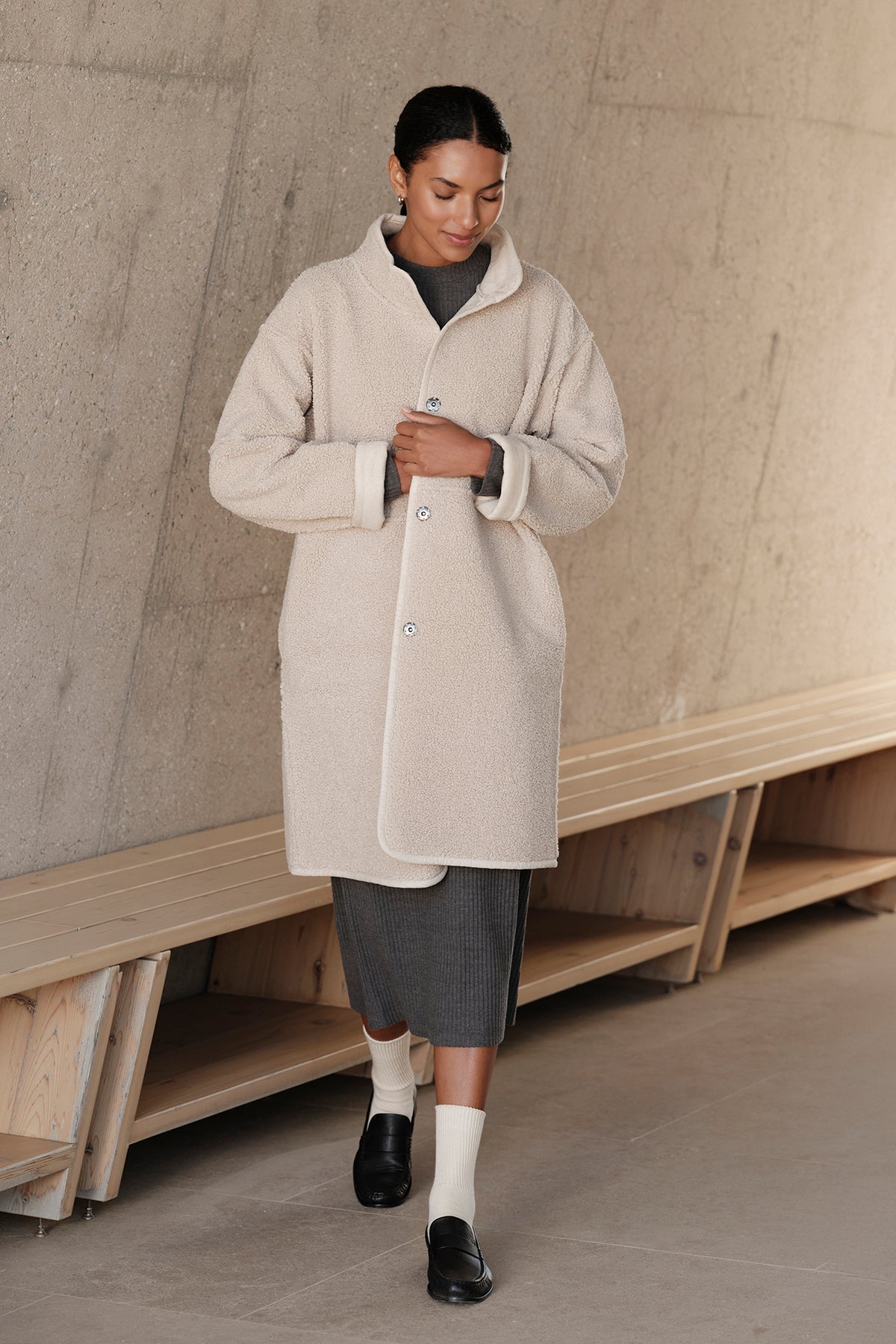   A person in a cream CARA LUXE SHERPA REVERSIBLE JACKET by Velvet by Graham & Spencer walks indoors, wearing a gray dress and black shoes. The setting includes a wooden bench and a concrete wall. 