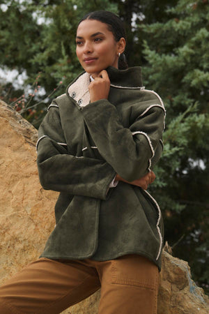 A person wearing the ALBANY LUXE SHERPA REVERSIBLE JACKET by Velvet by Graham & Spencer, featuring eco-friendly faux sherpa lining, stands outdoors, leaning against a rock with trees in the background.