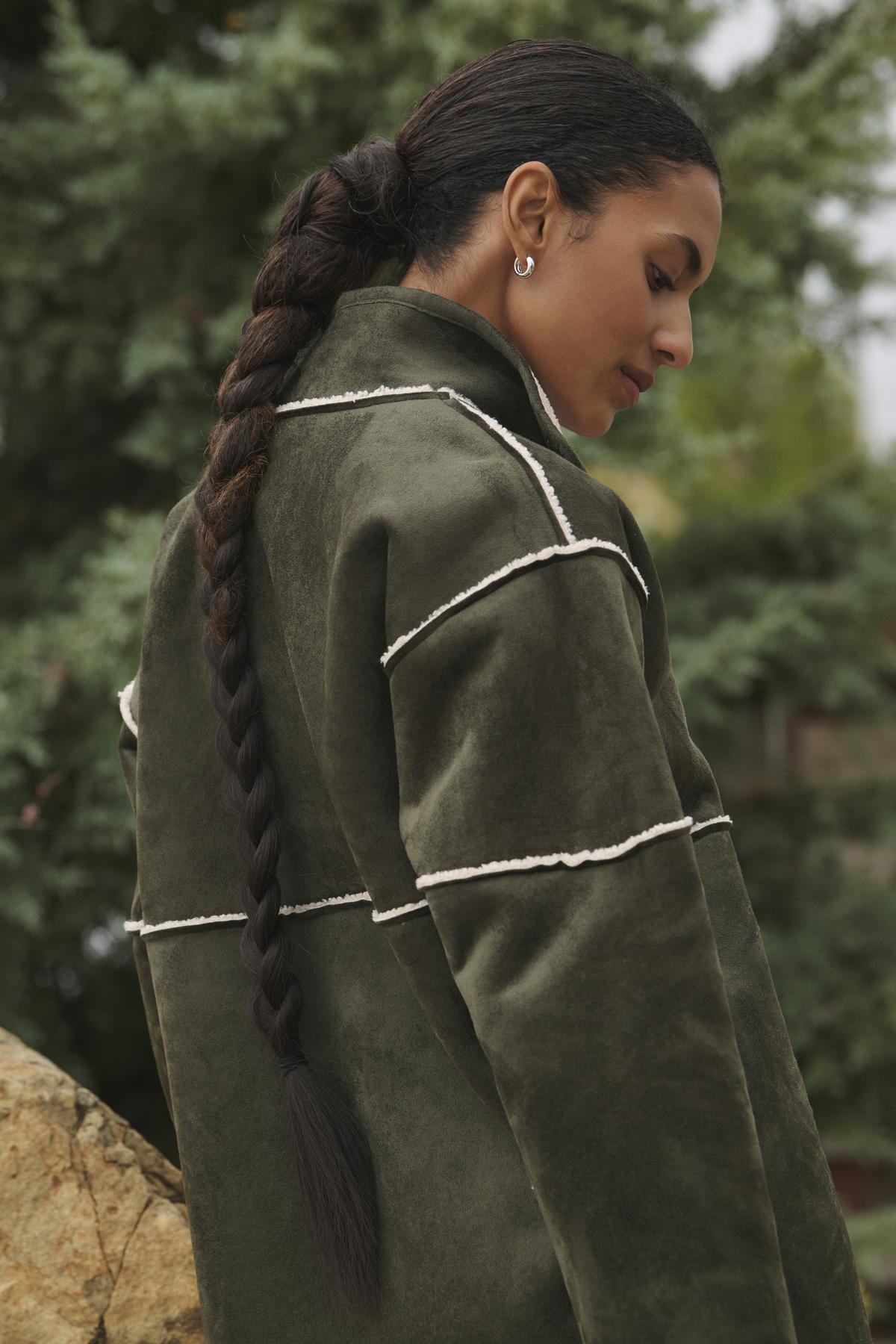 An individual with a lengthy braided ponytail is outdoors, glancing down while wearing the Velvet by Graham & Spencer's eco-friendly ALBANY LUXE SHERPA REVERSIBLE JACKET in olive green.-38219215175873