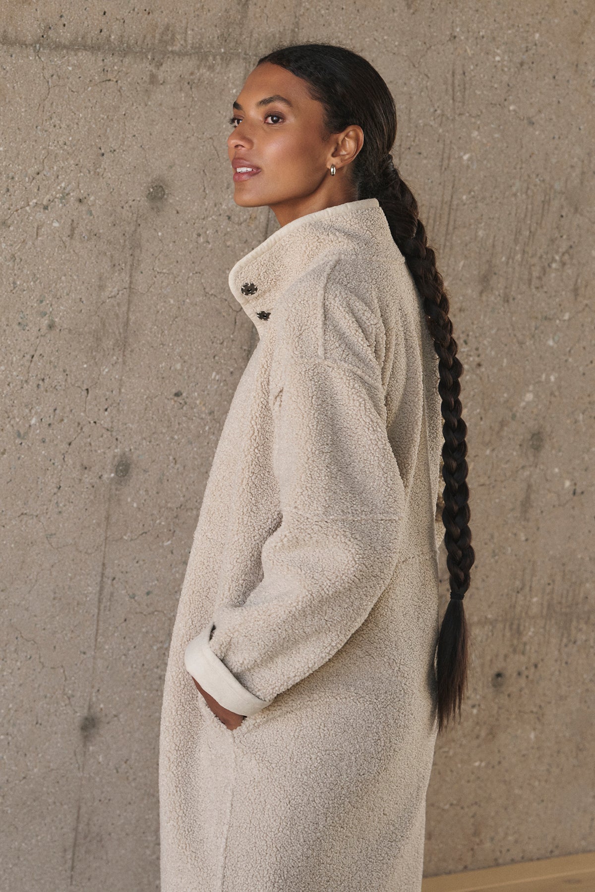   Individual with long braided hair wearing a light-colored, textured Cara Luxe Sherpa Reversible Jacket by Velvet by Graham & Spencer, standing against a concrete wall. 