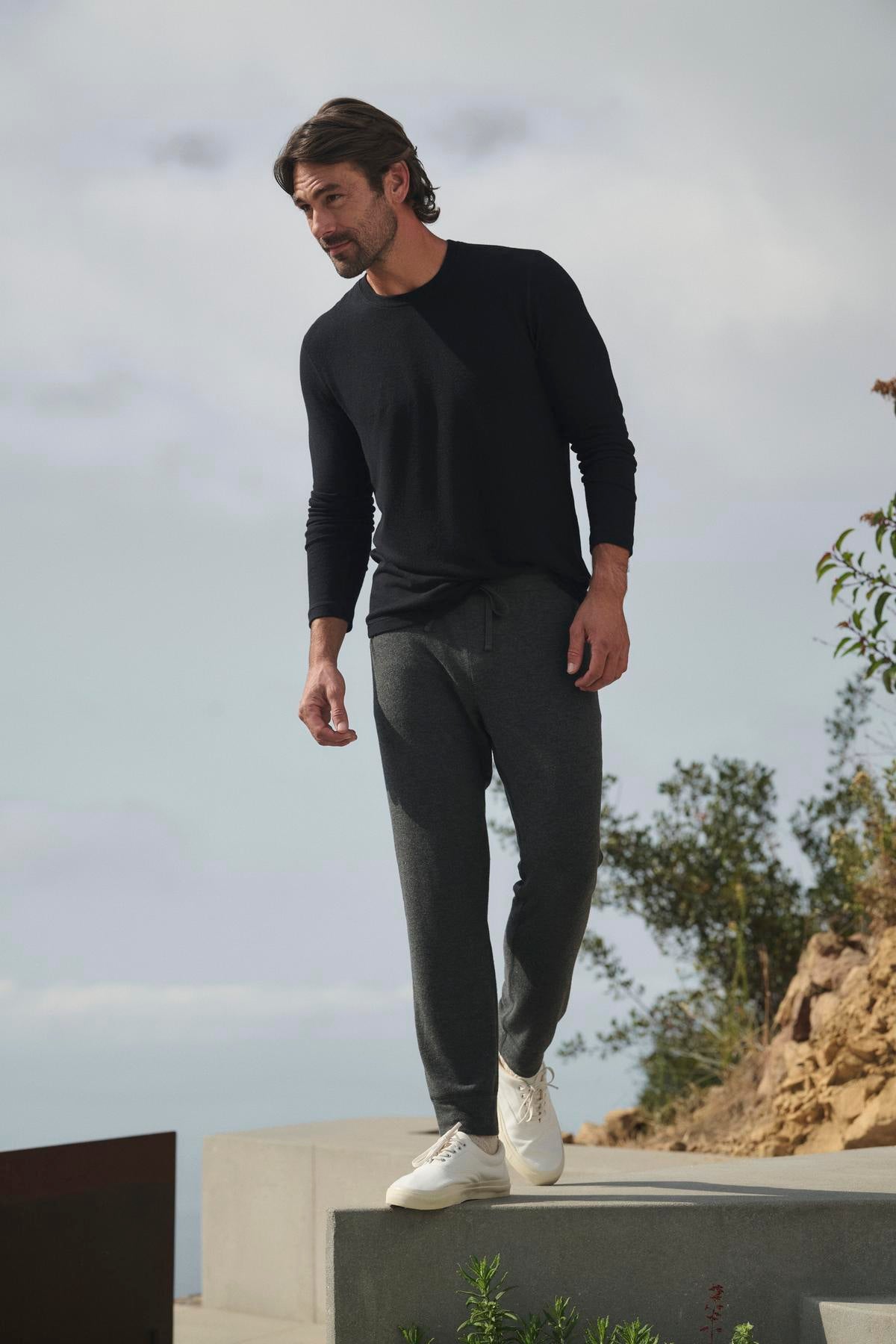 A man in a black long-sleeve shirt and the ZEALAND SWEATPANT by Velvet by Graham & Spencer strolls outdoors on a concrete path, with greenery and sky creating a serene backdrop.-38282318676161