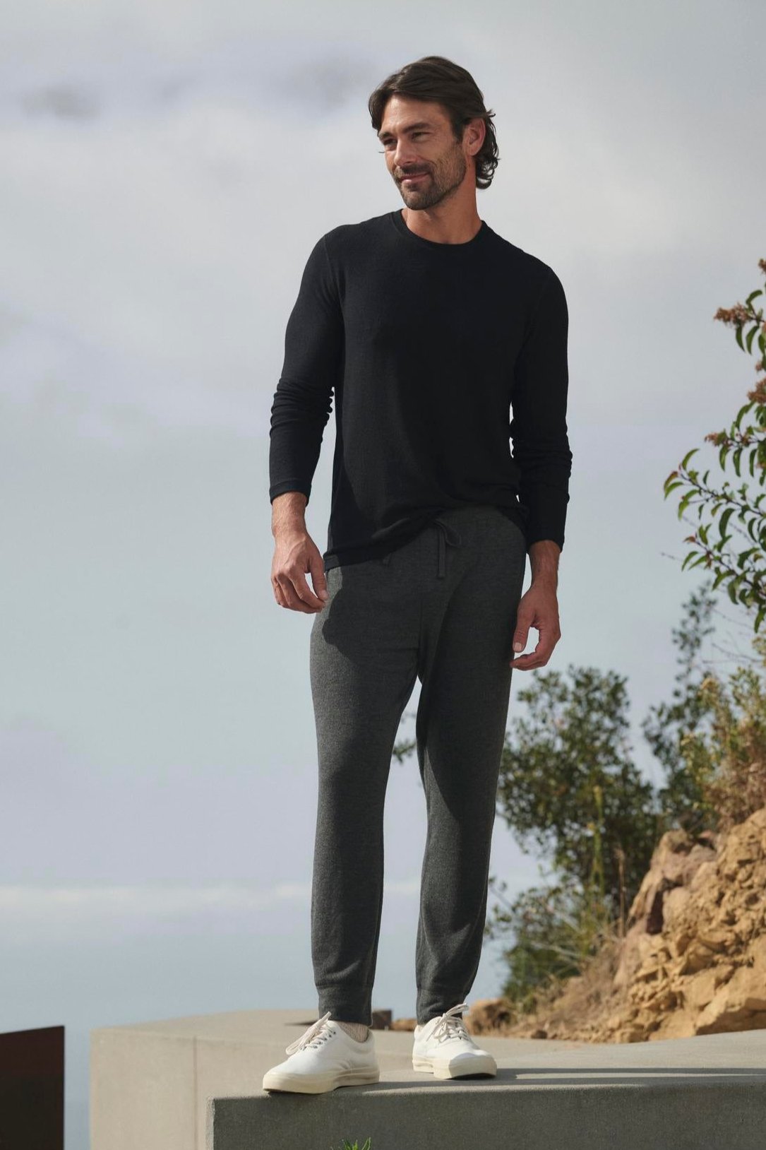   Wearing the ultra-soft BECKER CREW by Velvet by Graham & Spencer, a man in cozy black long-sleeve jersey material stands on a concrete platform outdoors. The gray pants complementing his outfit are set against lush plants and a cloudy sky, creating the perfect backdrop for his daily wear. 