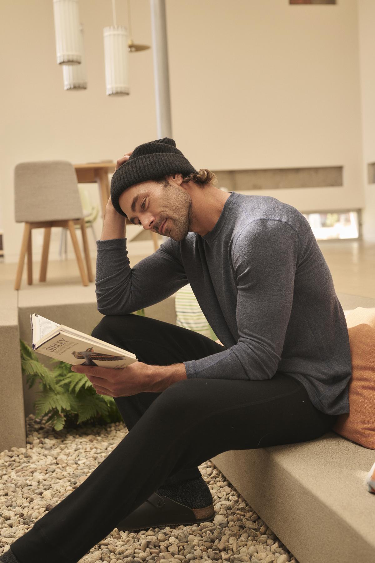 A person wearing a beanie, long-sleeve shirt, and ZEALAND SWEATPANT by Velvet by Graham & Spencer sits on a bench reading a book in a modern, light-filled room.-38282318840001