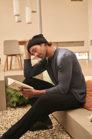 A person wearing a beanie, long-sleeve shirt, and ZEALAND SWEATPANT by Velvet by Graham & Spencer sits on a bench reading a book in a modern, light-filled room.