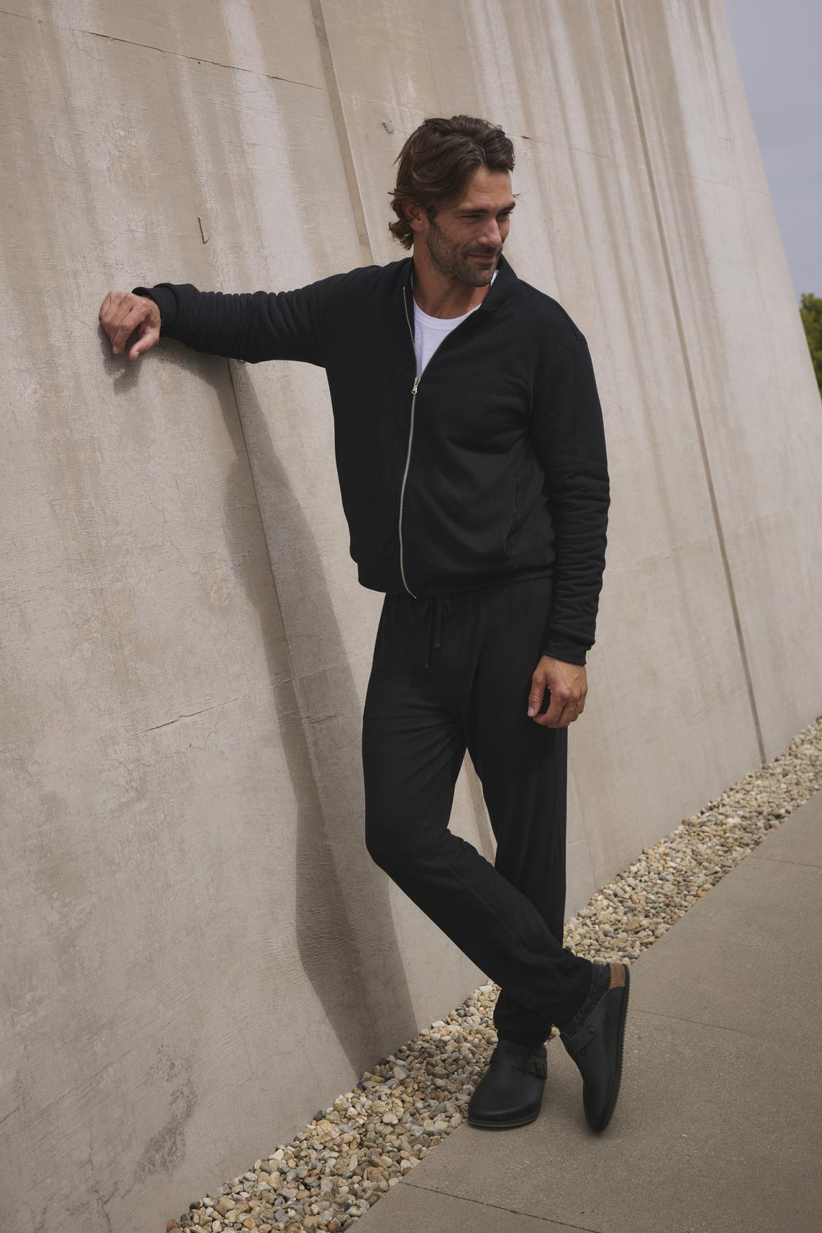 Dressed in a black ensemble featuring the ZEALAND SWEATPANT by Velvet by Graham & Spencer, he leans casually with his right hand against a concrete wall while standing on a pebble-lined path.-38282318708929