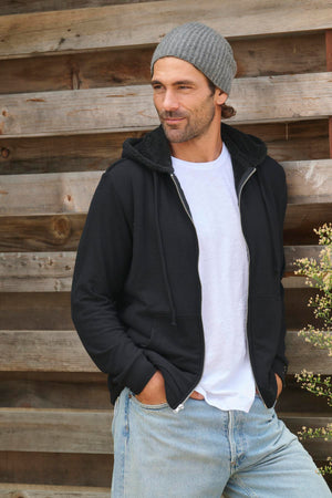 A person wearing a gray beanie, the SALVADORE HOODIE by Velvet by Graham & Spencer in ultra-soft, cozy jersey black, and a white shirt stands with hands in pockets in front of a wooden wall.