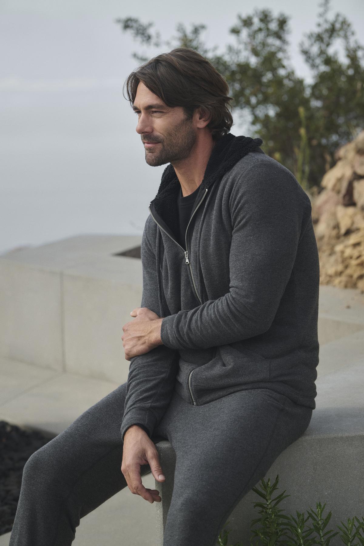   A man dressed in ZEALAND SWEATPANT by Velvet by Graham & Spencer sits outdoors on a concrete ledge with greenery in the background, wearing a gray hoodie and glancing to the side. 