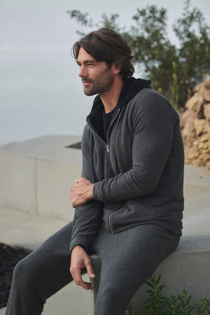 A man wearing the SALVADORE HOODIE by Velvet by Graham & Spencer, designed with ultra-soft cozy jersey fabric, sits on a stone ledge outdoors with a neutral expression, framed by plants in the background.