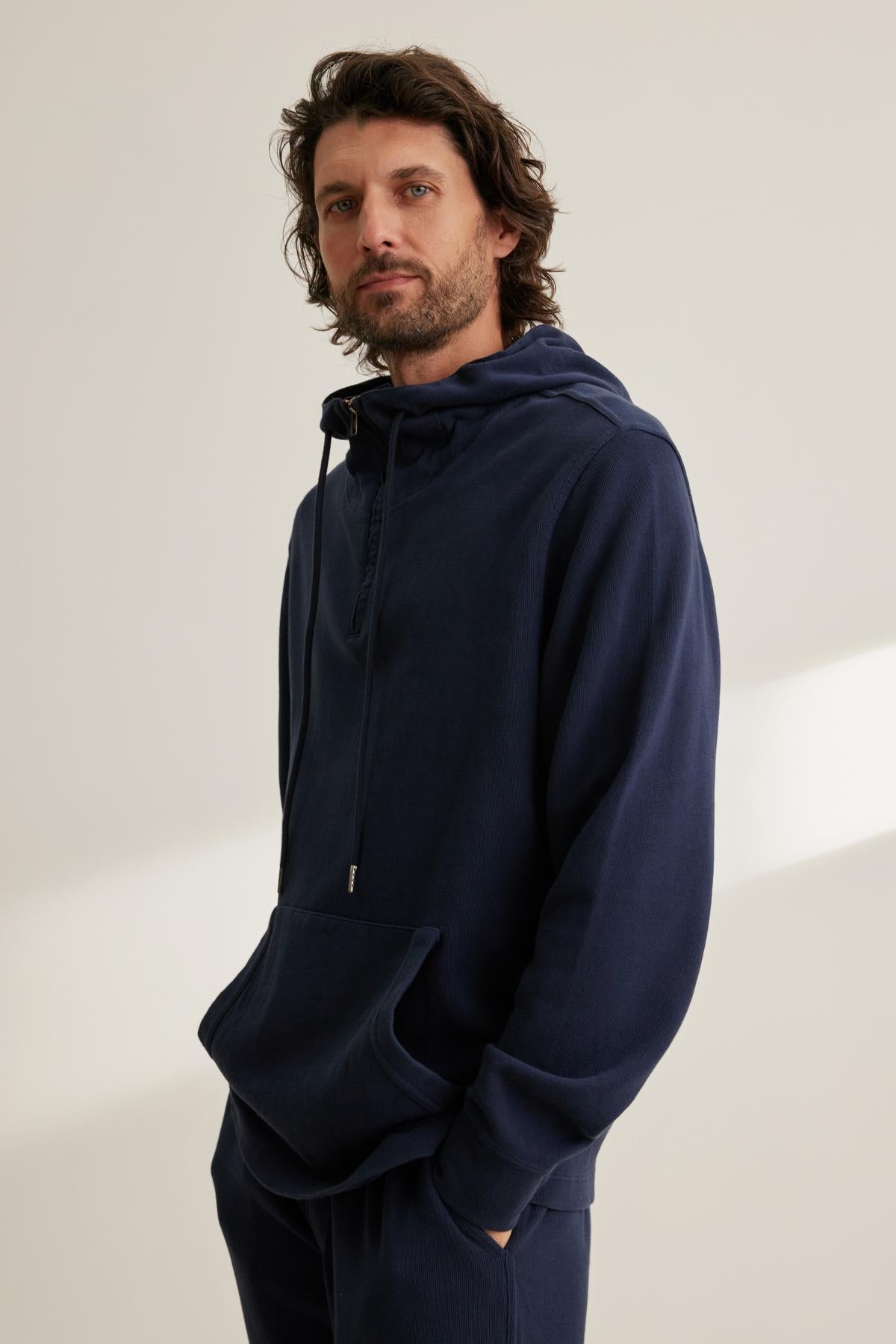   A man with wavy hair is wearing a Velvet by Graham & Spencer MORTY HOODIE in dark blue, complete with matching pants, standing against a light-colored background. 