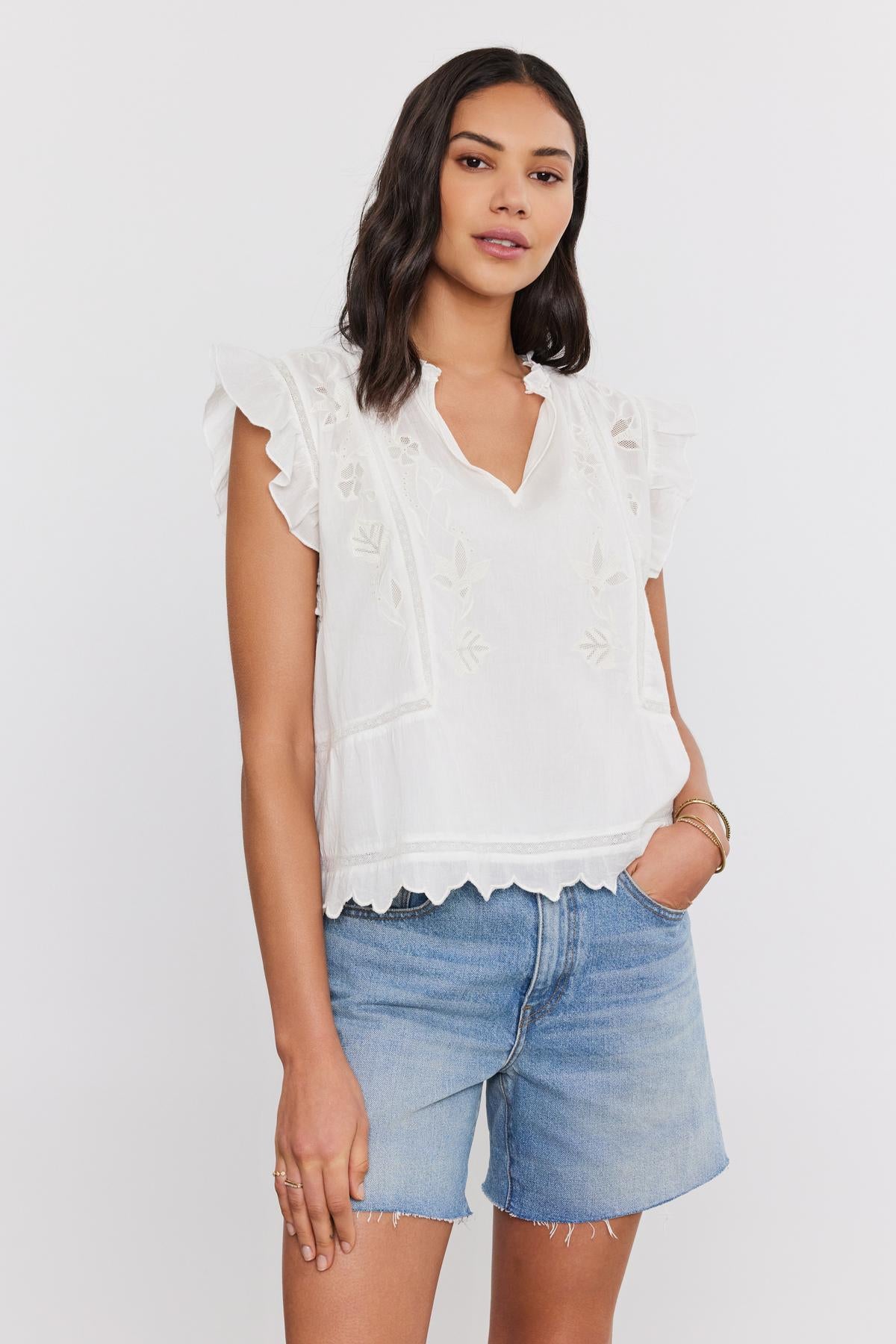 A woman in a white CHARLENE TOP with cotton embroidery and denim shorts, standing against a plain background. (Brand: Velvet by Graham & Spencer)-36830326751425