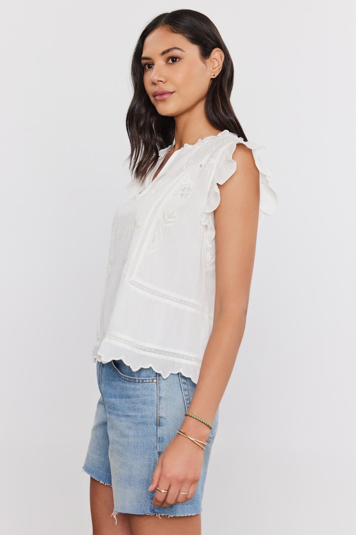   A woman wearing a white CHARLENE TOP with cotton embroidery and denim shorts, standing with a slight smile, facing towards the camera. Brand: Velvet by Graham & Spencer 