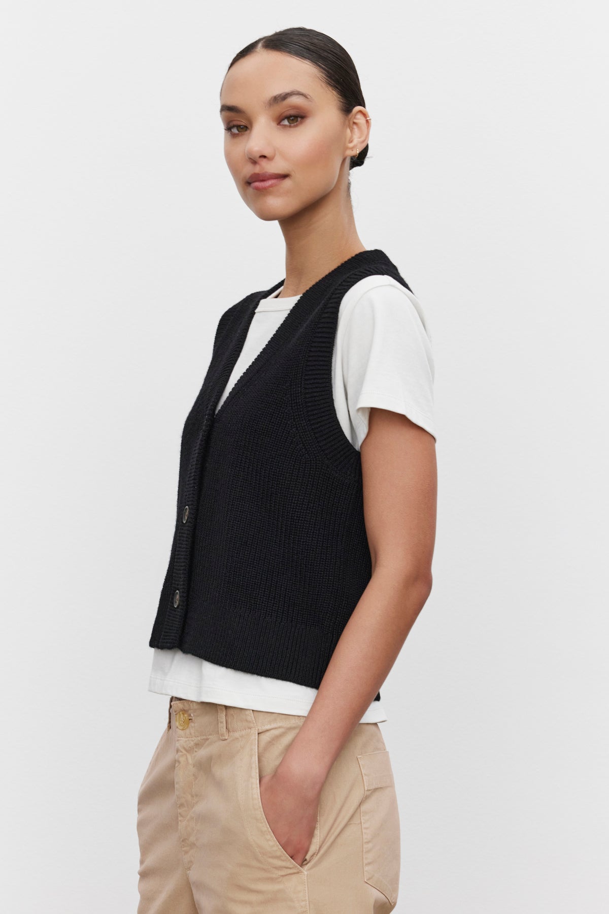   A person wearing the LU SWEATER VEST by Velvet by Graham & Spencer over a white t-shirt and beige pants, standing against a plain white background. 