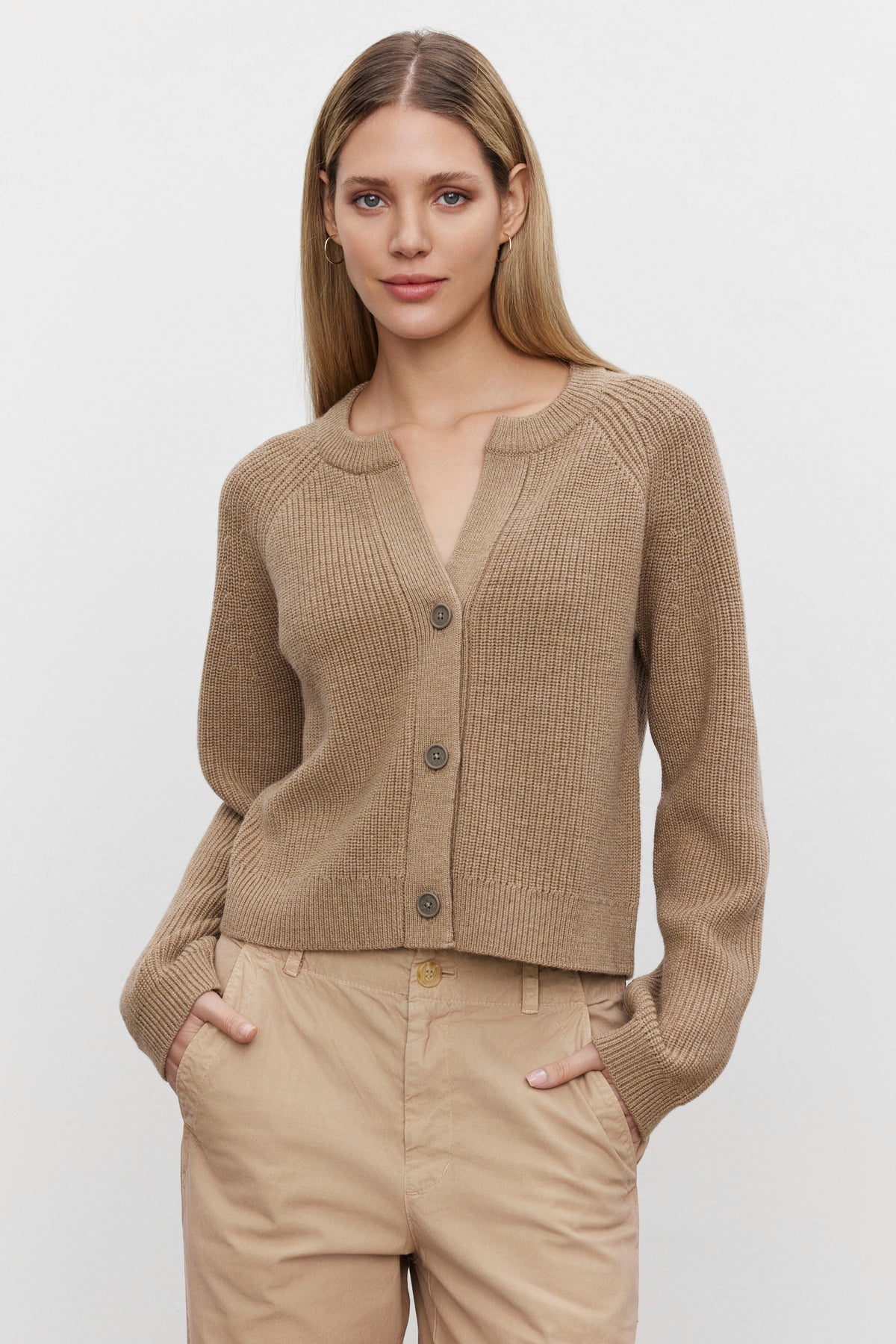 A woman with long blonde hair wears the SHAYLA CARDIGAN from Velvet by Graham & Spencer and beige pants, standing with her hands in her pockets against a neutral background.-37676908413121