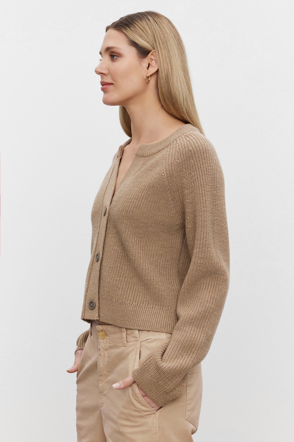 A woman with long blond hair, dressed in the SHAYLA CARDIGAN by Velvet by Graham & Spencer and tan pants, stands looking to the left against a plain white background.-37676908445889