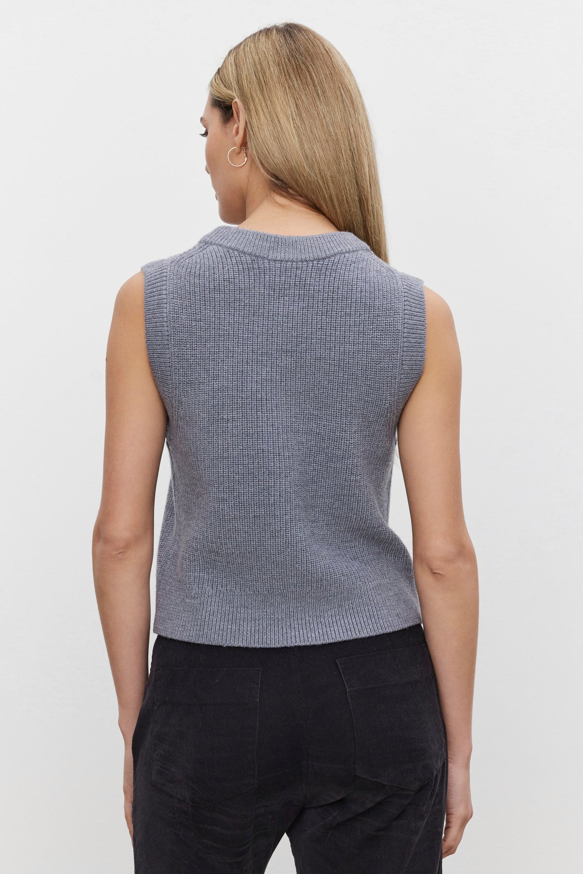   Back view of a person with long hair wearing a sleeveless LU SWEATER VEST by Velvet by Graham & Spencer, featuring a gray knit texture and a v-neckline, paired with dark pants. 