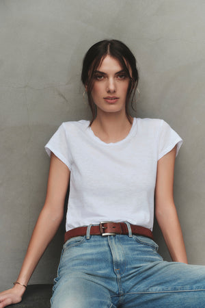 A woman with dark hair is wearing a white Velvet by Graham & Spencer ODELIA TEE with a classic crew neckline, blue jeans, and a brown belt, sitting against a gray wall.