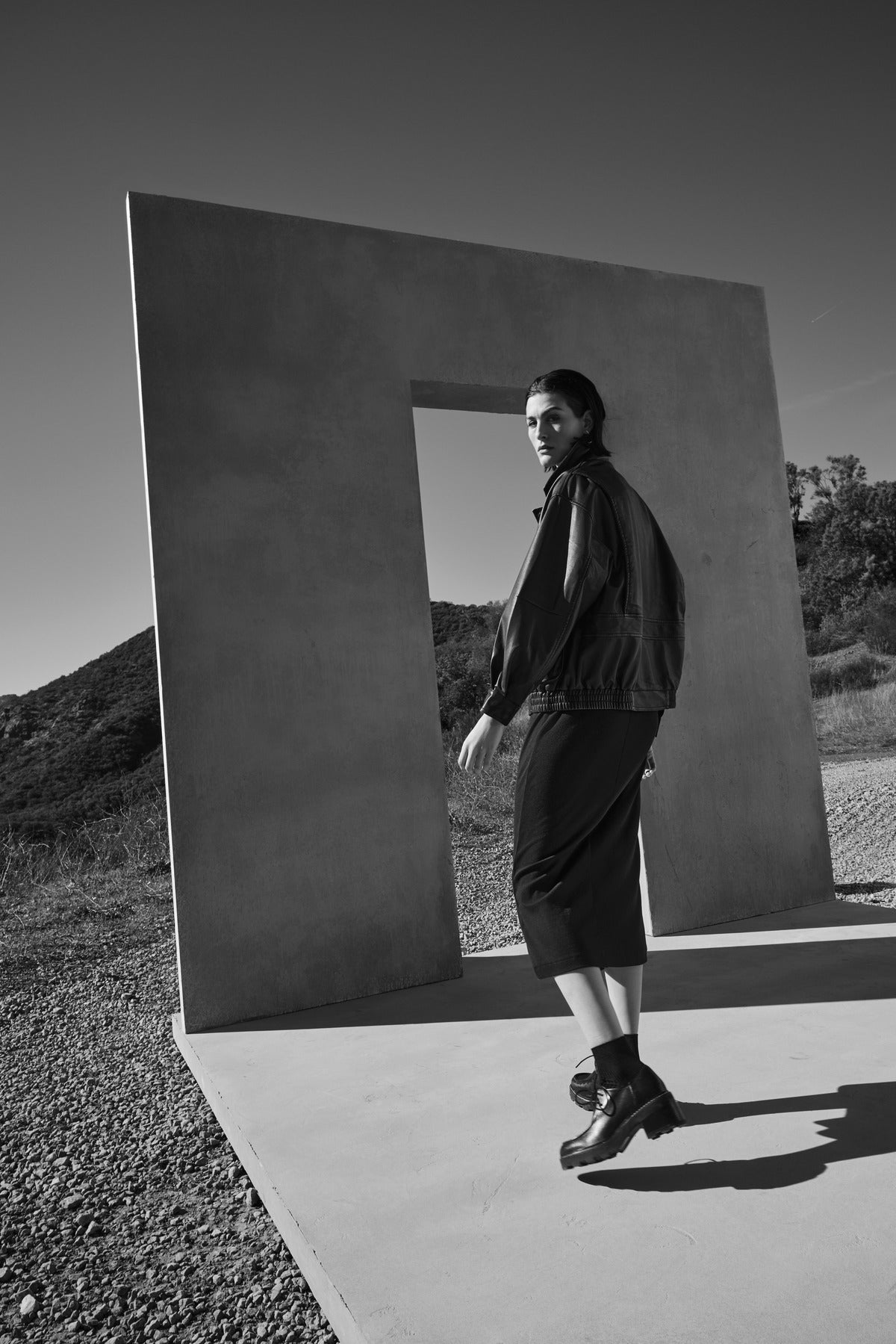   Black and white photo of a person wearing the CALIFORNIA FAUX LEATHER JACKET by Velvet by Jenny Graham, standing near a large rectangular concrete structure with a centered opening, on a gravel surface with hills in the background. 