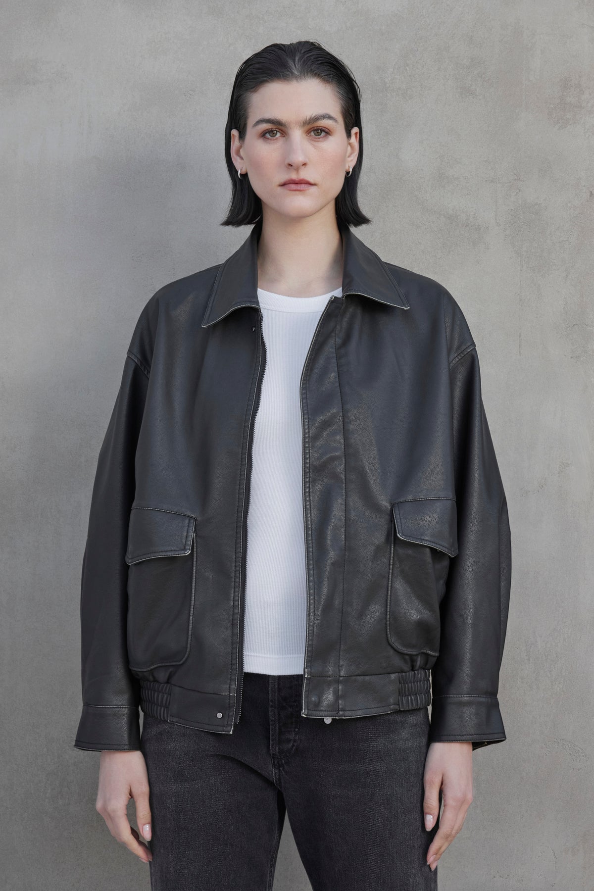   A person with short dark hair is wearing a CALIFORNIA FAUX LEATHER JACKET from Velvet by Jenny Graham over a white shirt and dark pants, standing against a plain concrete wall. 