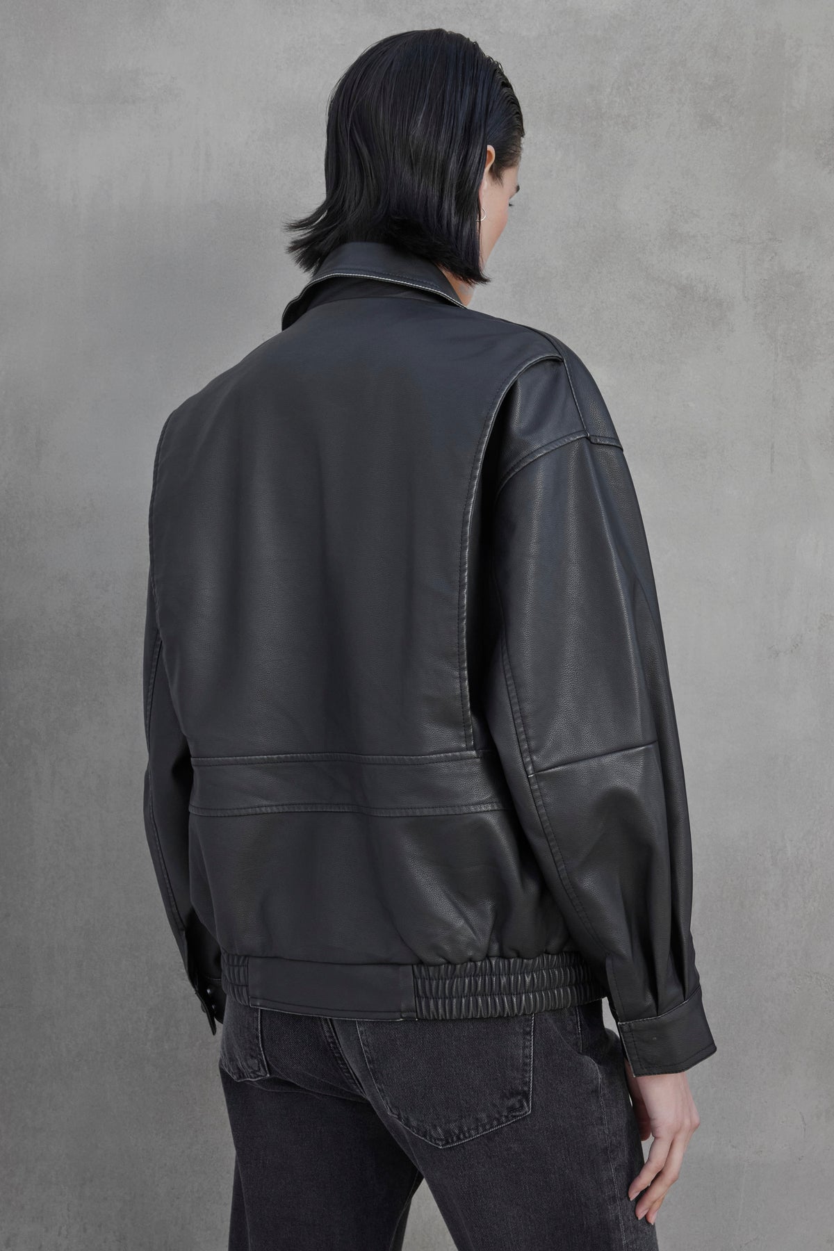 A person with short dark hair, seen from the back, is wearing a CALIFORNIA FAUX LEATHER JACKET by Velvet by Jenny Graham and dark jeans, standing in front of a textured gray wall.-37659910308033