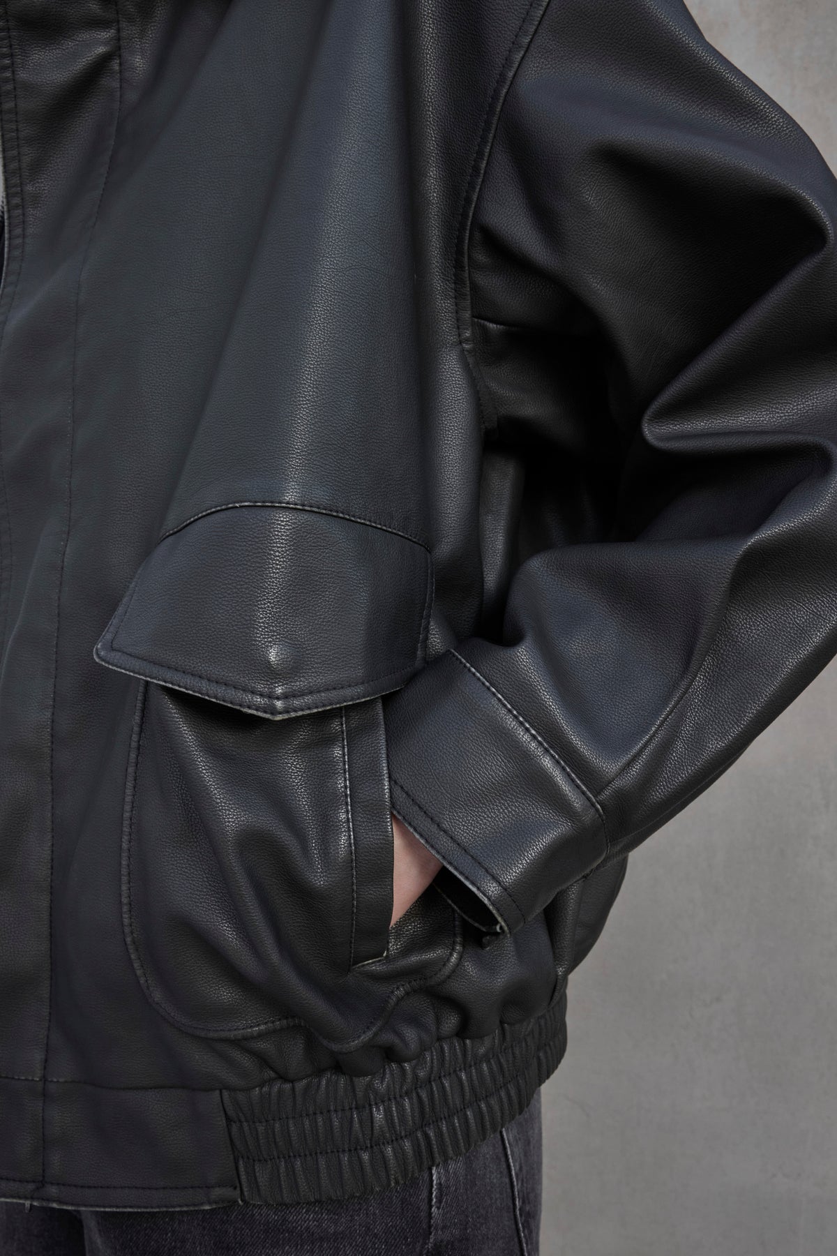   Close-up of a person wearing the CALIFORNIA FAUX LEATHER JACKET by Velvet by Jenny Graham, featuring a loose fit with their hand in a pocket. The black jacket boasts a side flap pocket and elastic cuffs. 