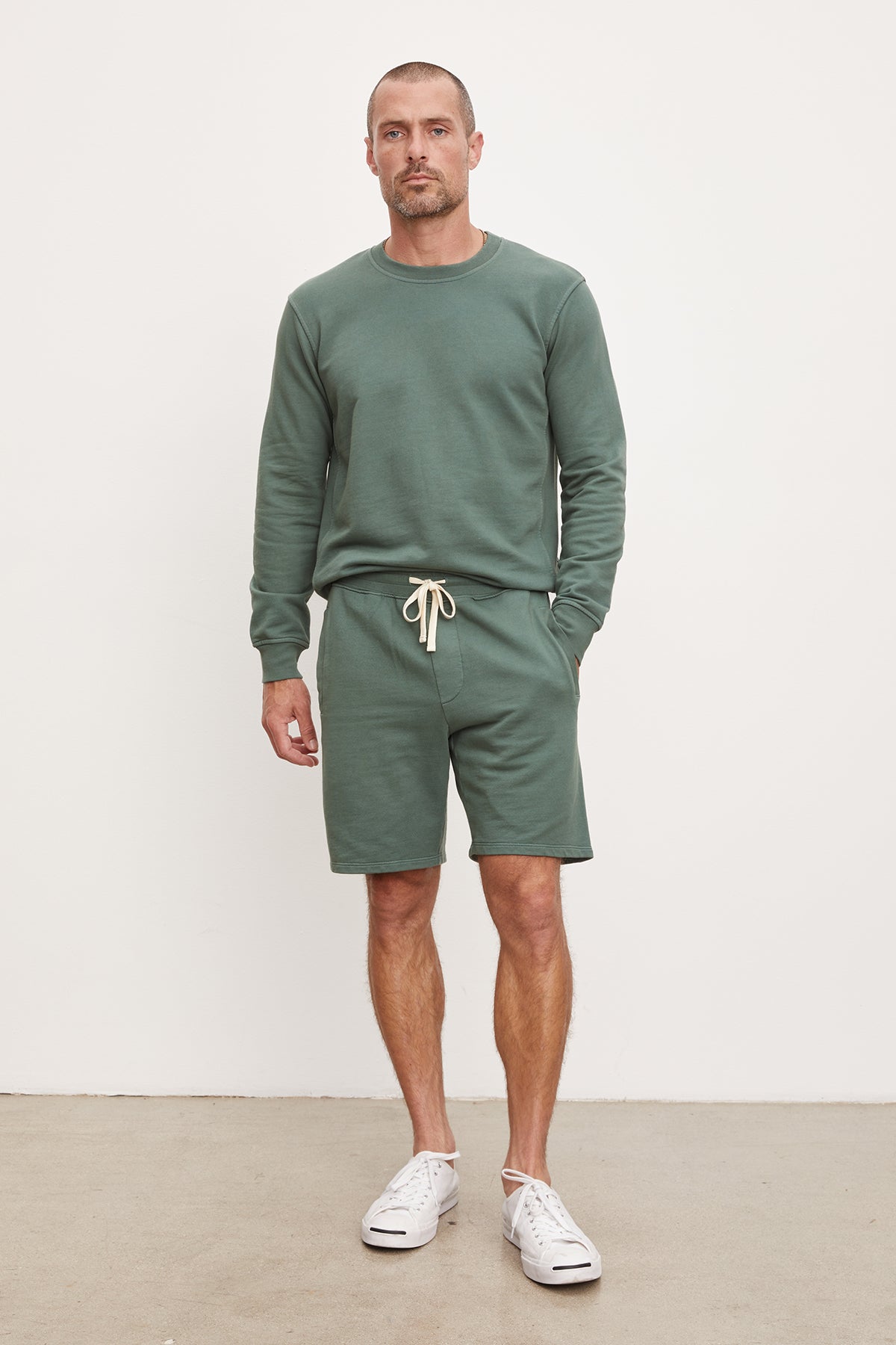   A man stands against a plain background, wearing a green sweatshirt, French terry Beckett shorts from Velvet by Graham & Spencer, and white sneakers. 