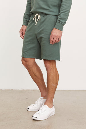 A man wearing a green sweatshirt and matching Velvet by Graham & Spencer Beckett shorts with white sneakers, cropped to show from the chest down.