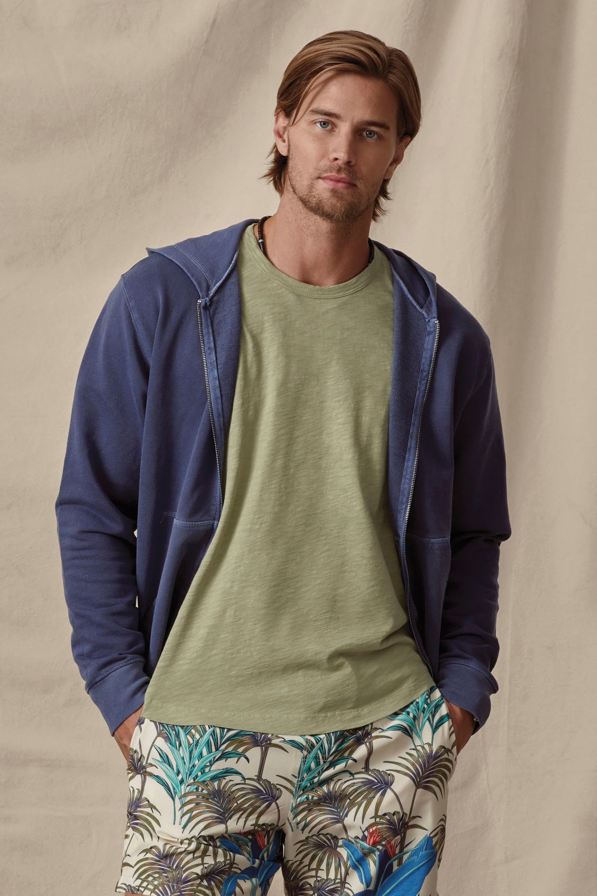   A man with shoulder-length hair wearing a Velvet by Graham & Spencer Vincent Hoodie, green t-shirt, and tropical print pants, standing against a beige backdrop. 