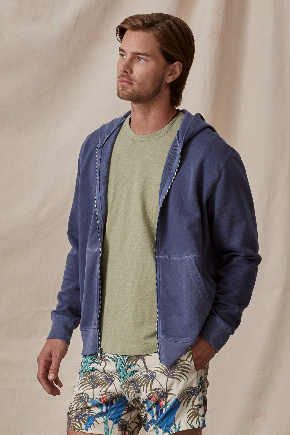   Man in a casual outfit featuring a Velvet by Graham & Spencer VINCENT HOODIE, green t-shirt, and tropical print shorts, standing against a beige backdrop. 