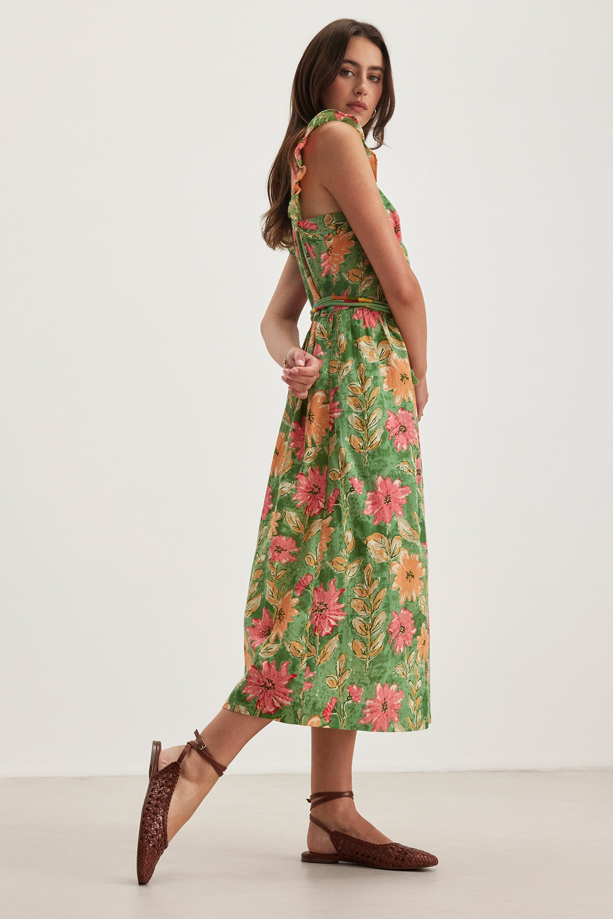   A woman in the ELARIA DRESS by Velvet by Graham & Spencer, featuring a green floral print with flutter sleeves and a detachable braided belt, stands sideways against a plain background wearing brown shoes. 