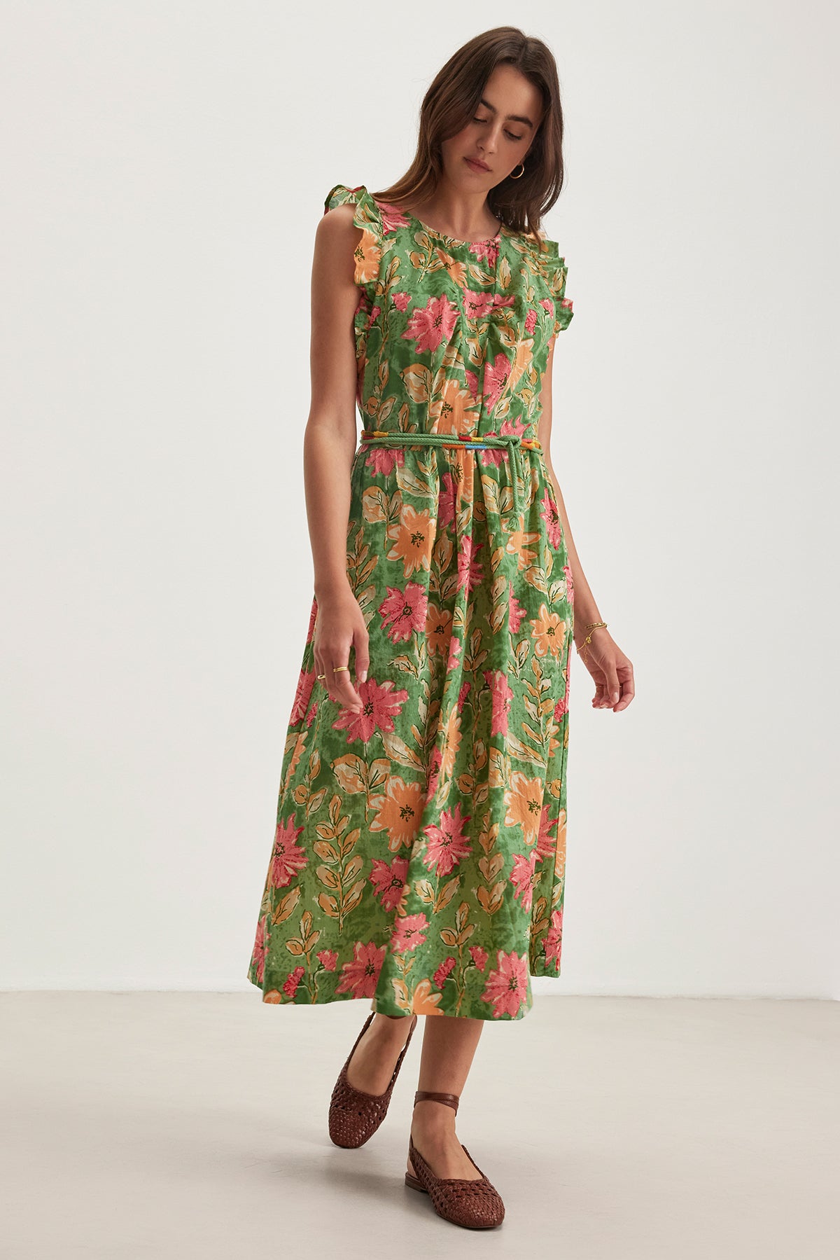 A woman with brown hair and woven brown shoes wears the ELARIA DRESS, a green cotton dress with pink and orange flowers and flutter sleeves by Velvet by Graham & Spencer, against a plain white background.-38662636765377