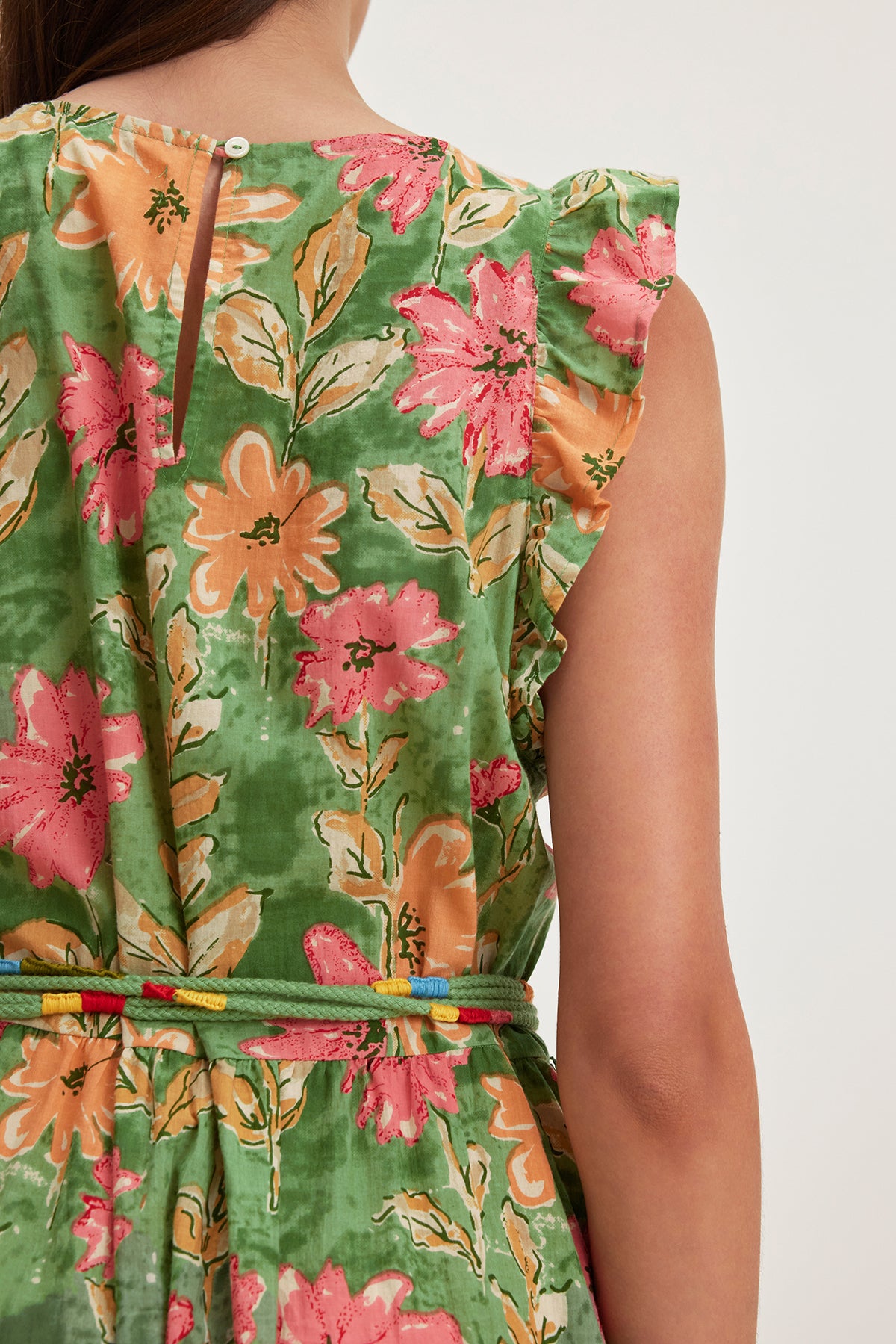   A person is seen from the back wearing the ELARIA DRESS by Velvet by Graham & Spencer, a green floral cotton dress with pink and orange flowers, featuring flutter sleeves and a detachable braided belt. 